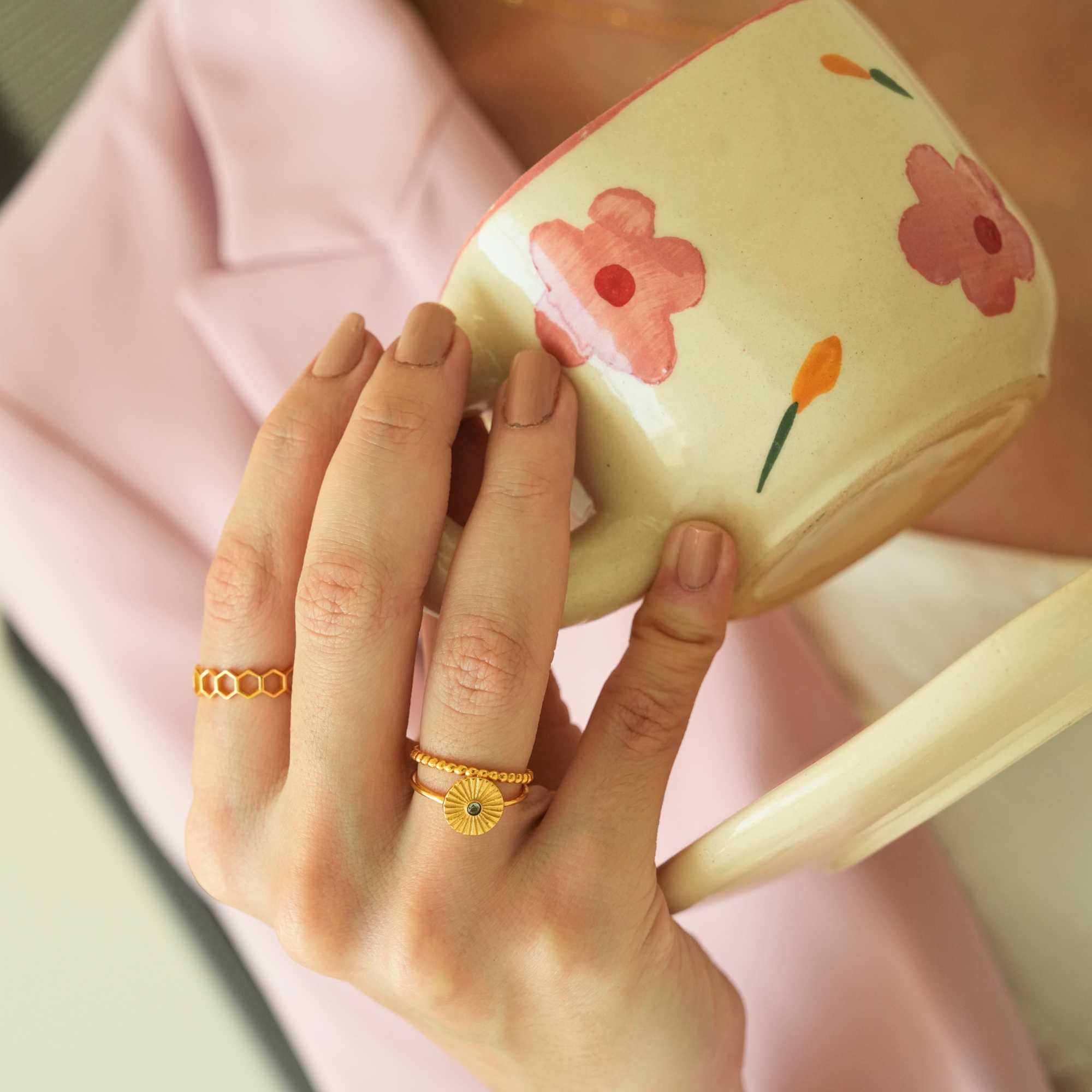 Angled lifestyle close-up of a hand holding a flower-patterned mug against a soft pink blazer backdrop, highlighting the textured stack of the Delicate Vintage Beaded Stacking Ring Gold and a sunburst ring, by Protsaah Zurich.