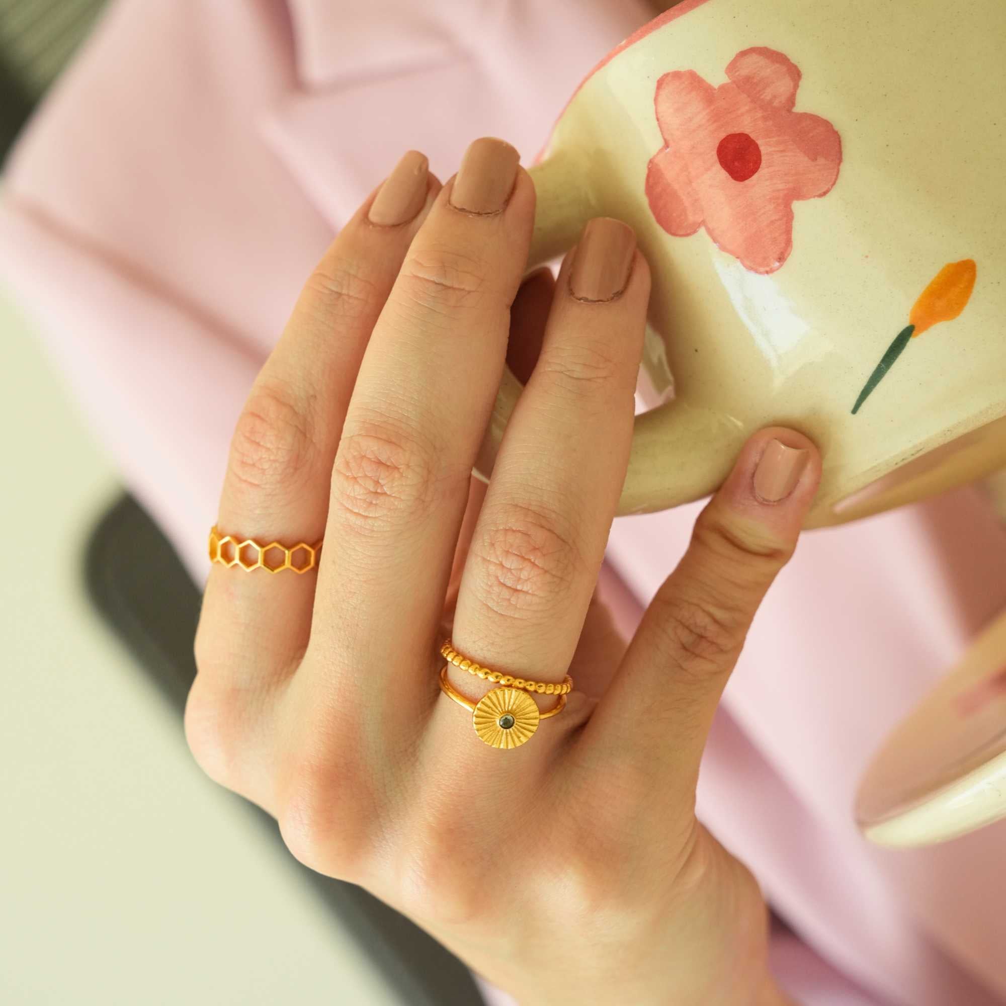 Close-up lifestyle shot of a model's hand gripping a hand-painted floral ceramic mug, showcasing the Delicate Vintage Beaded Stacking Ring Gold stacked beneath a sunburst signet ring, by Protsaah Zurich.