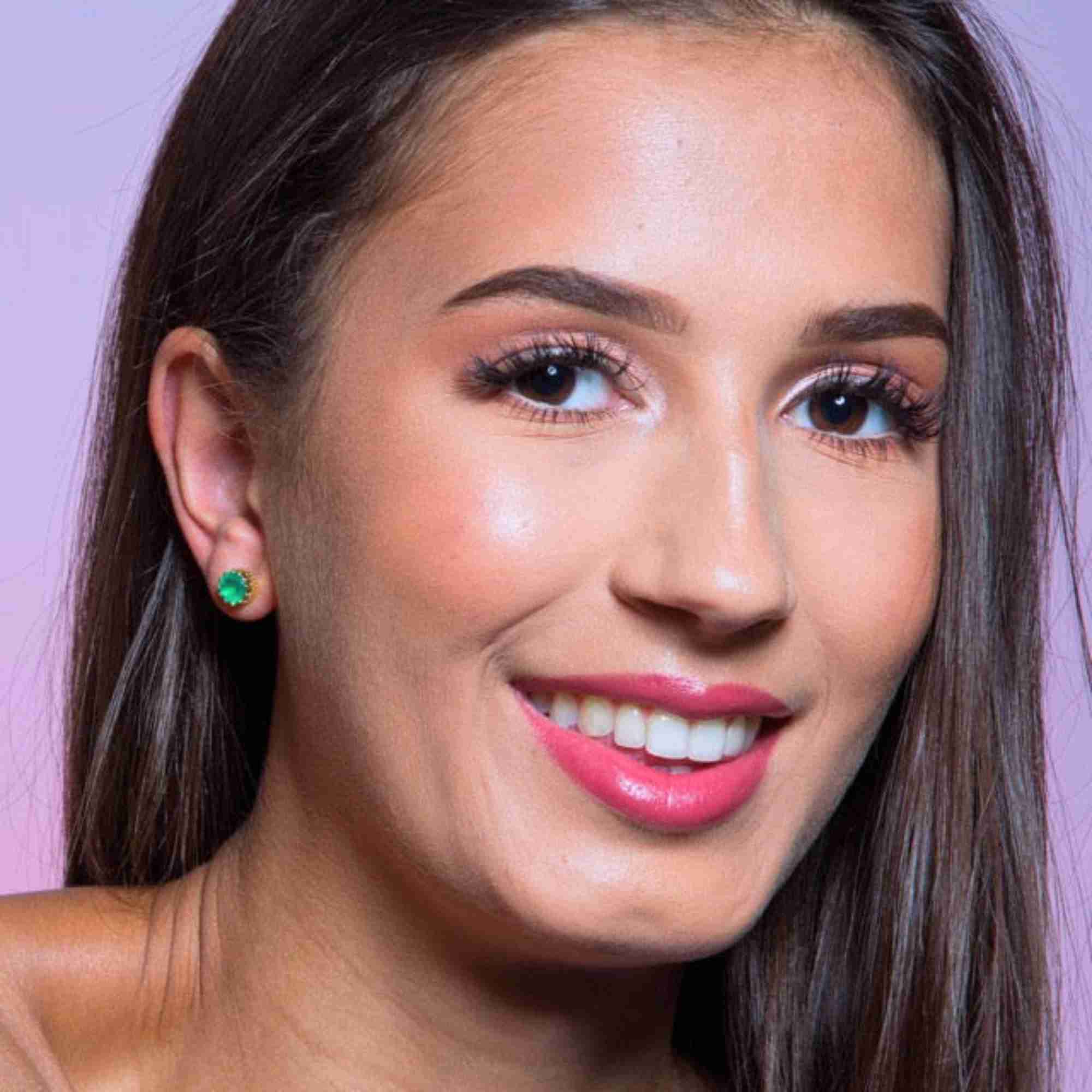 Editorial campaign picture named Dew Drops displaying Dew Drops Aqua Chalcedony Crown Stud Earrings Silver. Close-up portrait of a smiling female model with long dark brown hair and radiant pink lipstick, modeling a handcrafted round faceted light green-blue gemstone stud earring nested within a vintage crown setting on her earlobe. Shot against a soft pastel purple-pink background by Protsaah Zurich.
