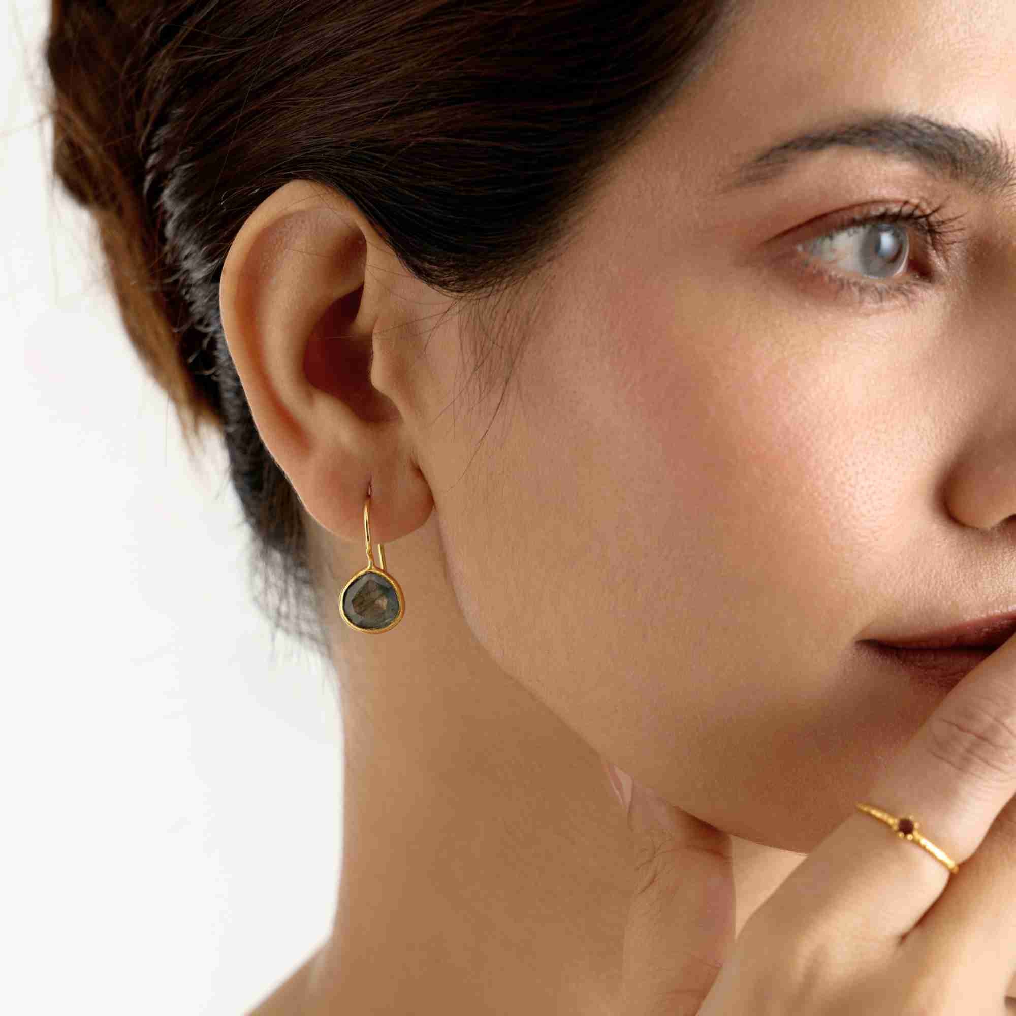 Studio lifestyle view of a model accessorized with the Dew Drops Labradorite Textured Drop Earrings Gold, referencing file name Dew_Drops_Collections_2.jpg. She brings a hand up toward her mouth, revealing a thin gold stacker ring on her finger while drawing focus up to the matching labradorite hook earring catching the light.