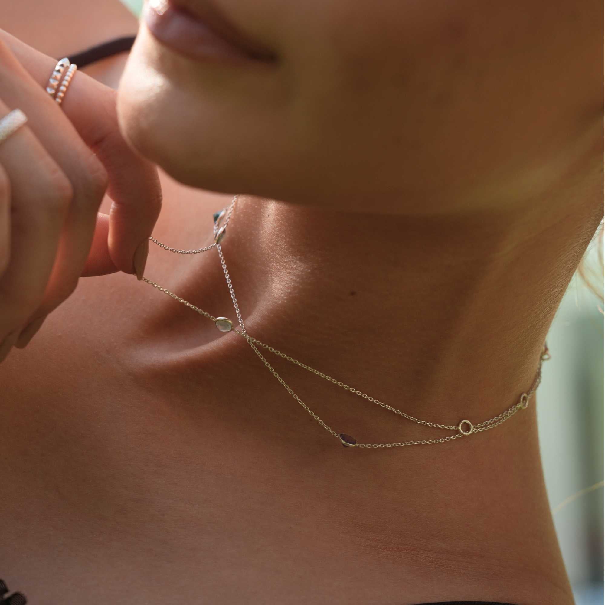 Close-up detail of a model adjusting the fine 92.5 sterling silver cable chain of the Dew Drops multi-gemstone necklace, emphasizing its delicate station charms and lightweight fluid drape.