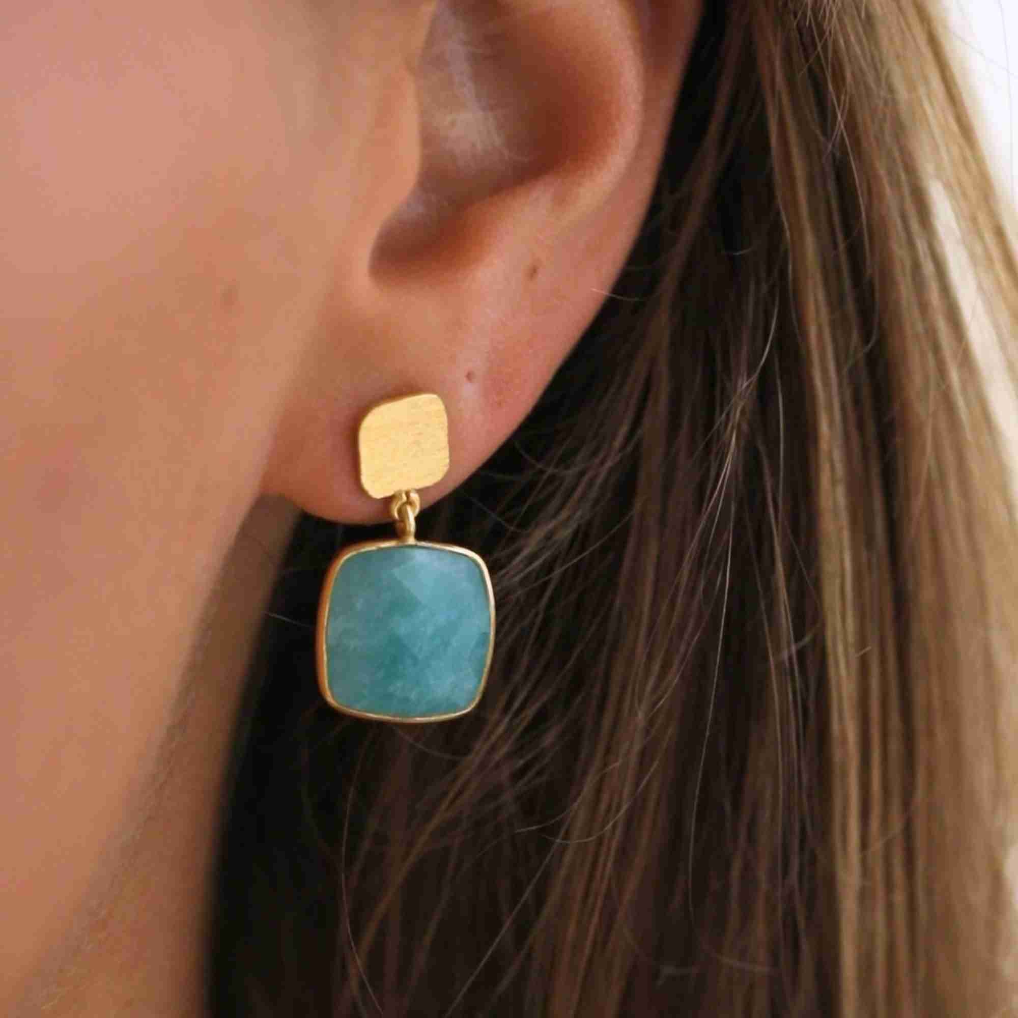 Detailed close-up lifestyle picture named Geo Glam focusing on a model's ear wearing a handcrafted 22K gold vermeil drop earring, featuring a square, cushion-cut translucent light turquoise-blue Amazonite gemstone suspended from a textured square gold post by Protsaah Zurich.
