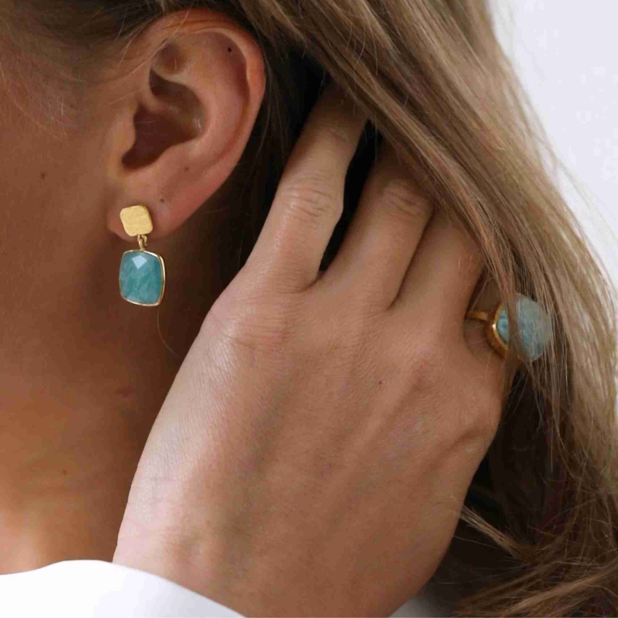 Lifestyle portrait campaign picture named Geo Glam showing a blonde model brushing back her hair with her hand to reveal her ear wearing the Cushion-Cut Amazonite Drop Earrings Gold, paired with a matching gold Amazonite gemstone ring on her finger by Protsaah Zurich.
