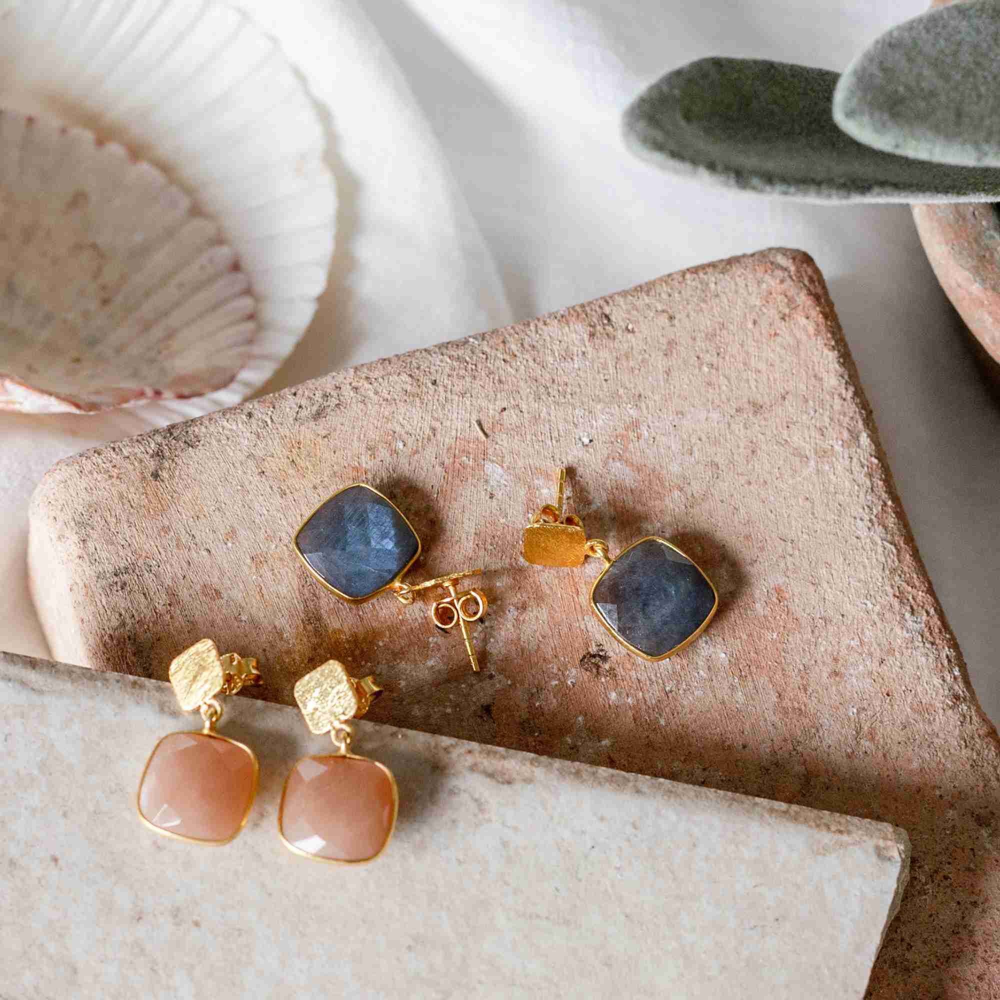 Lifestyle flatlay picture named Geo Glam displaying the Cushion-Cut Labradorite Drop Earrings Gold alongside peach moonstone drop earrings. The handcrafted 22K gold vermeil pairs are elegantly arranged on stacked rustic terracotta tiles next to a white seashell and green succulent leaves by Protsaah Zurich.