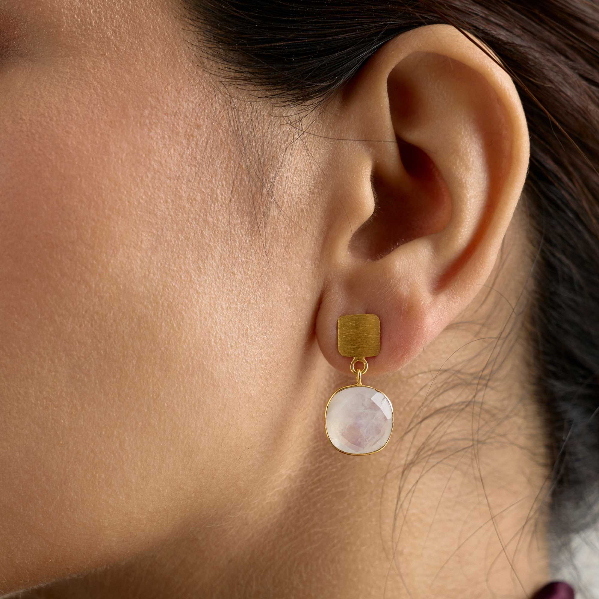 Extreme close-up lifestyle profile view focusing on a model's ear, highlighting the details of the Geo Glam Cushion-Cut Rainbow Moonstone Drop Earrings Gold. The shot clearly displays the fine texture on the square gold metal stud holding a shimmering, faceted white moonstone droplet.