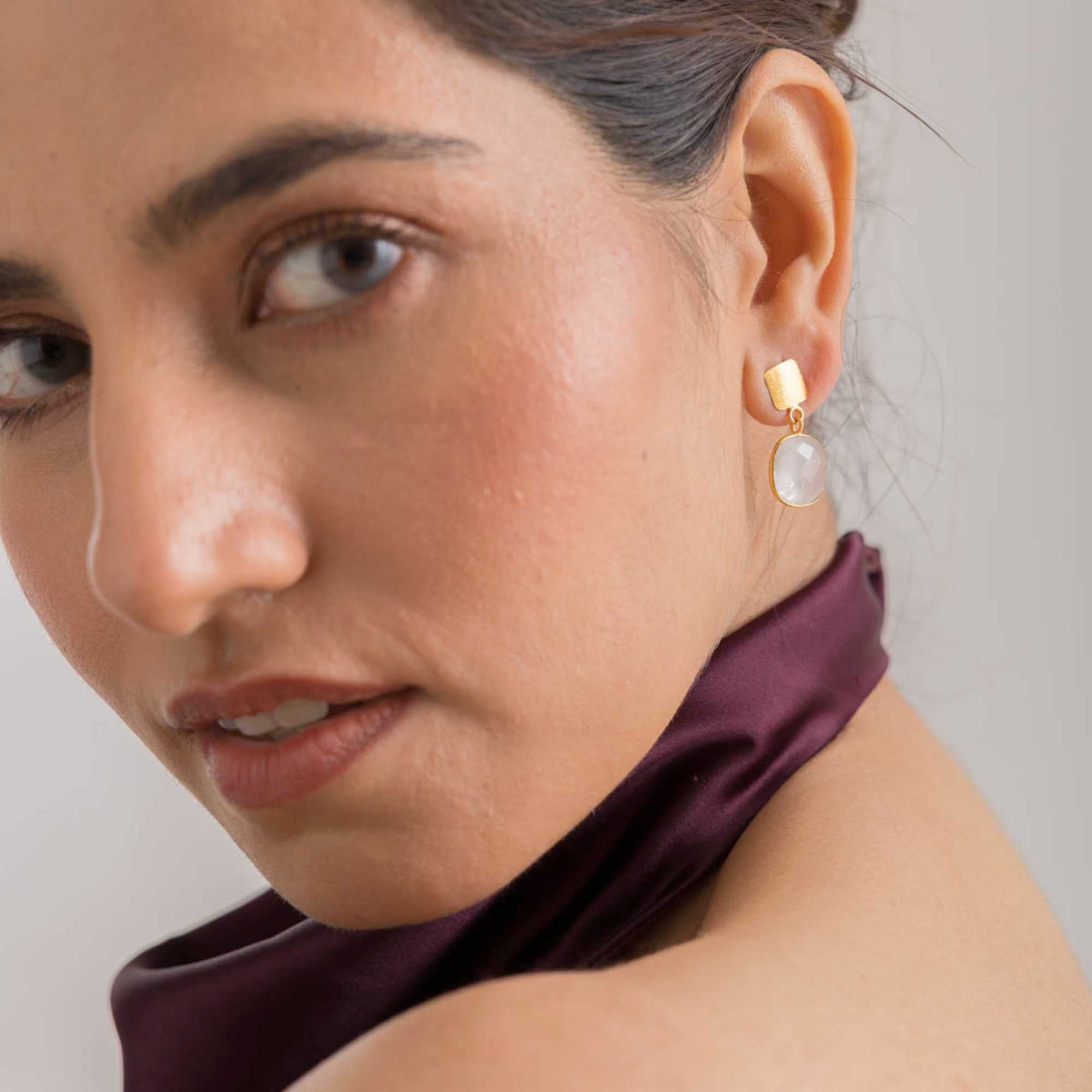 Angled lifestyle portrait of a model looking over her shoulder, showcasing the Geo Glam Cushion-Cut Rainbow Moonstone Drop Earrings Gold. She wears a plum-colored satin top while the earring captures the light with its small textured square gold stud and dangling translucent cushion-cut gemstone.