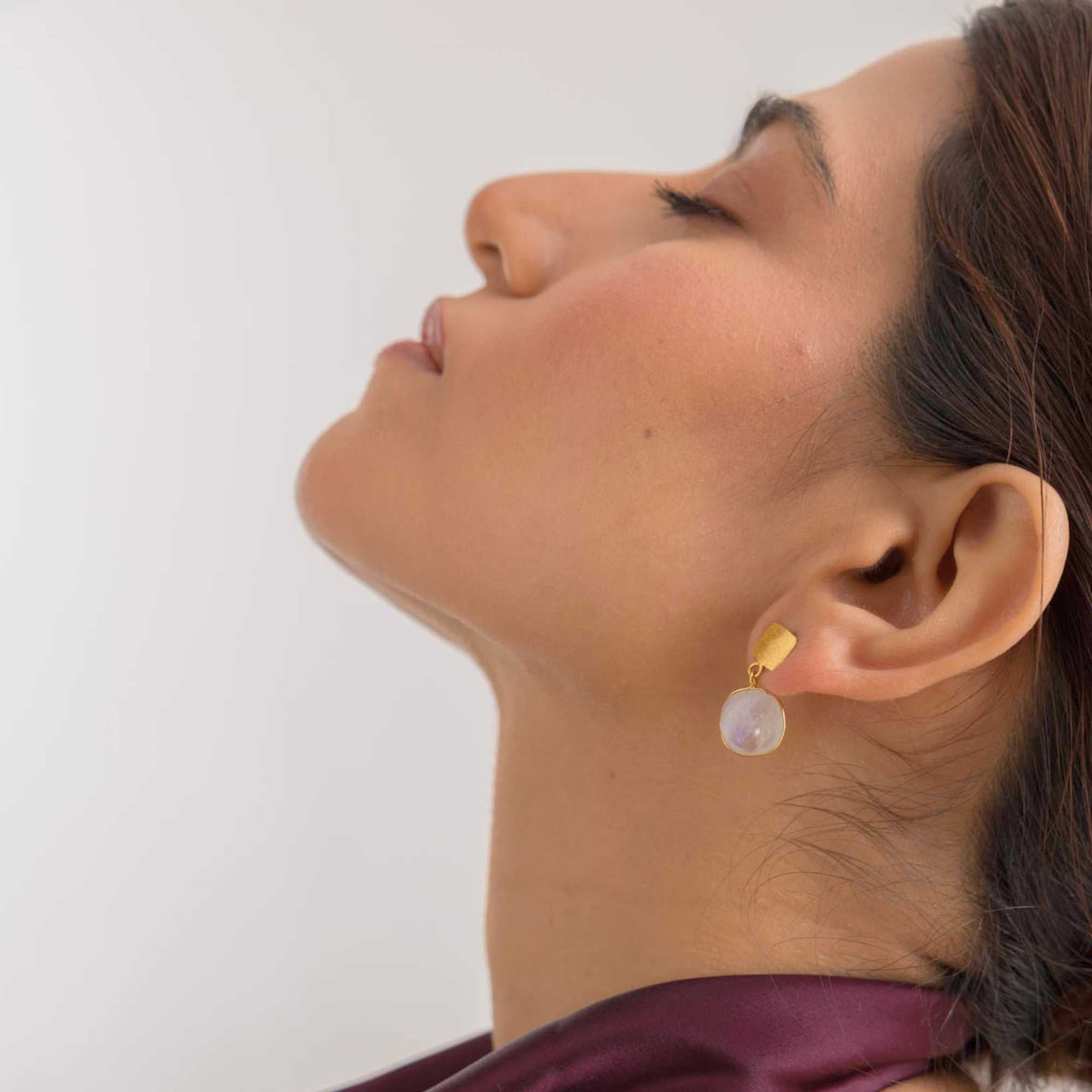 Lifestyle profile shot of a model tilting her head back with eyes closed, displaying the Geo Glam Cushion-Cut Rainbow Moonstone Drop Earrings Gold. Her elegant pose highlights the gentle drop of the gold square stud and translucent cushion-cut moonstone against her neckline.