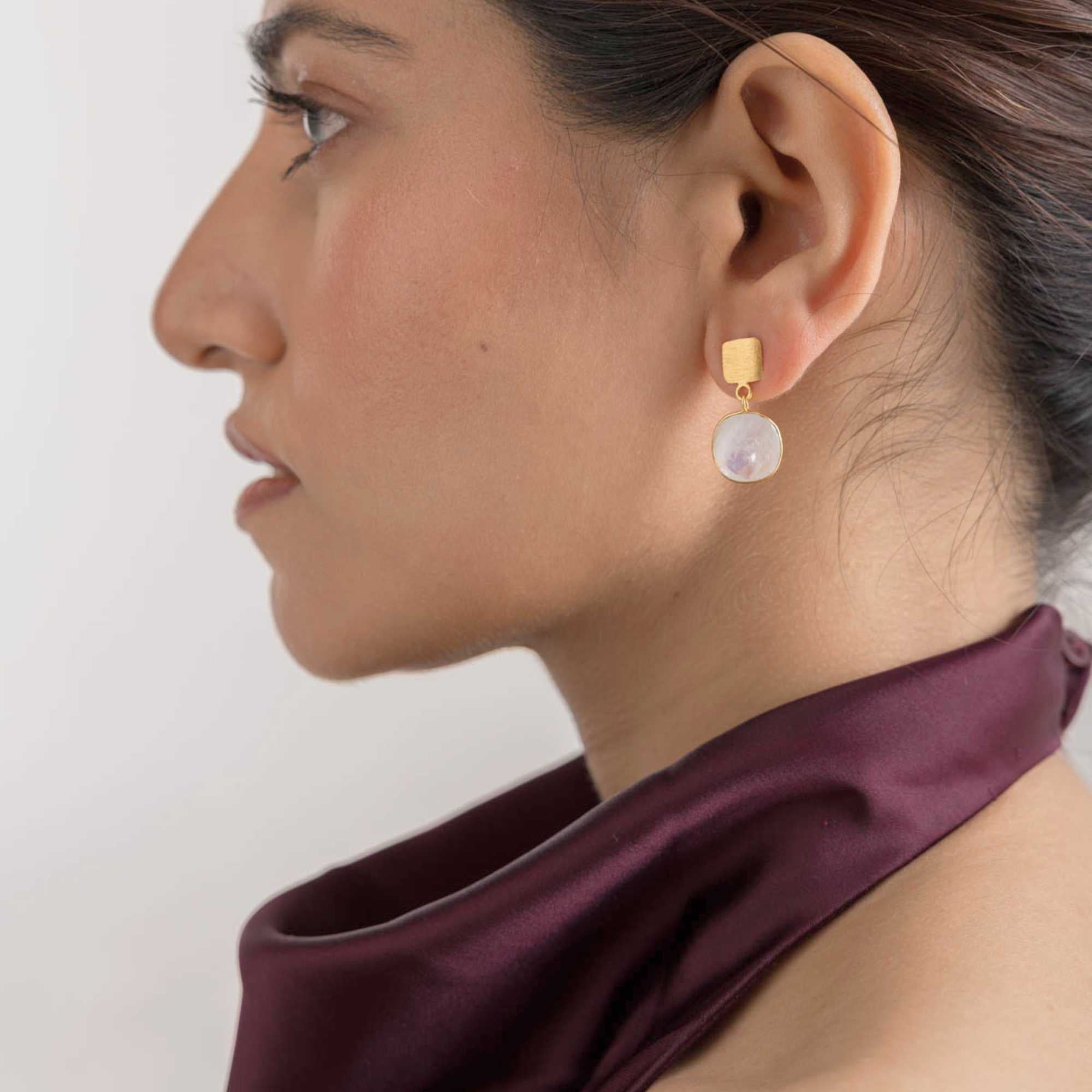 Classic profile lifestyle portrait of a model looking slightly to the side, showcasing the Geo Glam Cushion-Cut Rainbow Moonstone Drop Earrings Gold. The soft illumination emphasizes her facial features and the elegant shimmer of the geometric gold moonstone drop earring.