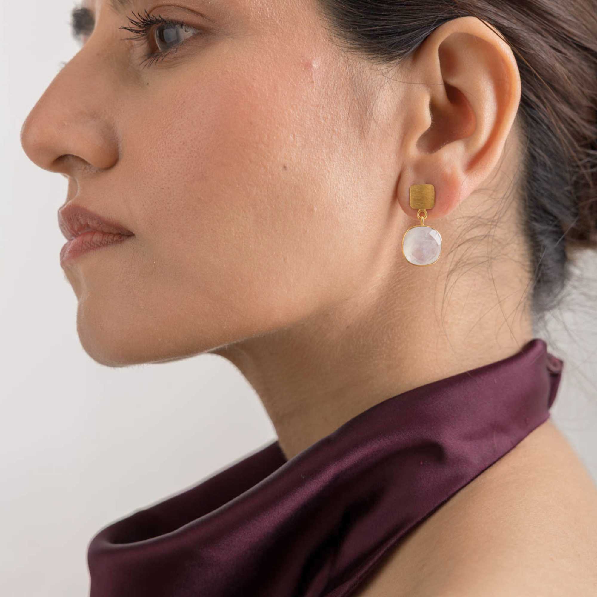 Three-quarter profile lifestyle view of a model looking ahead, featuring the Geo Glam Cushion-Cut Rainbow Moonstone Drop Earrings Gold. The gold-plated square base and dangling translucent moonstone bring a modern geometric flair to her deep purple satin ensemble.