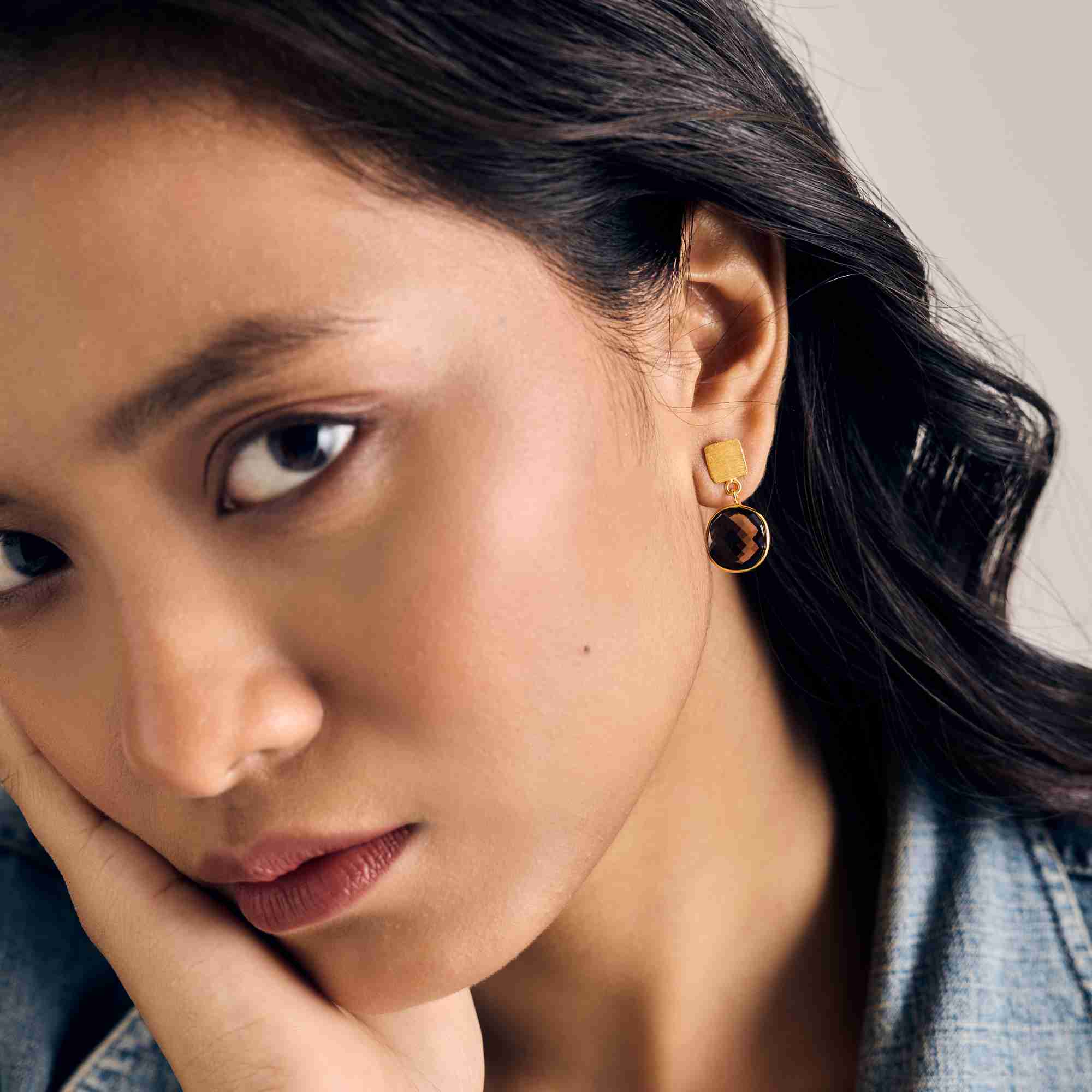 Close-up portrait campaign picture named Geo Glam showing a dark-haired model in a denim jacket resting her hand on her chin, showcasing the Cushion-Cut Smoky Quartz Drop Earrings Gold in her earlobe by Protsaah Zurich.