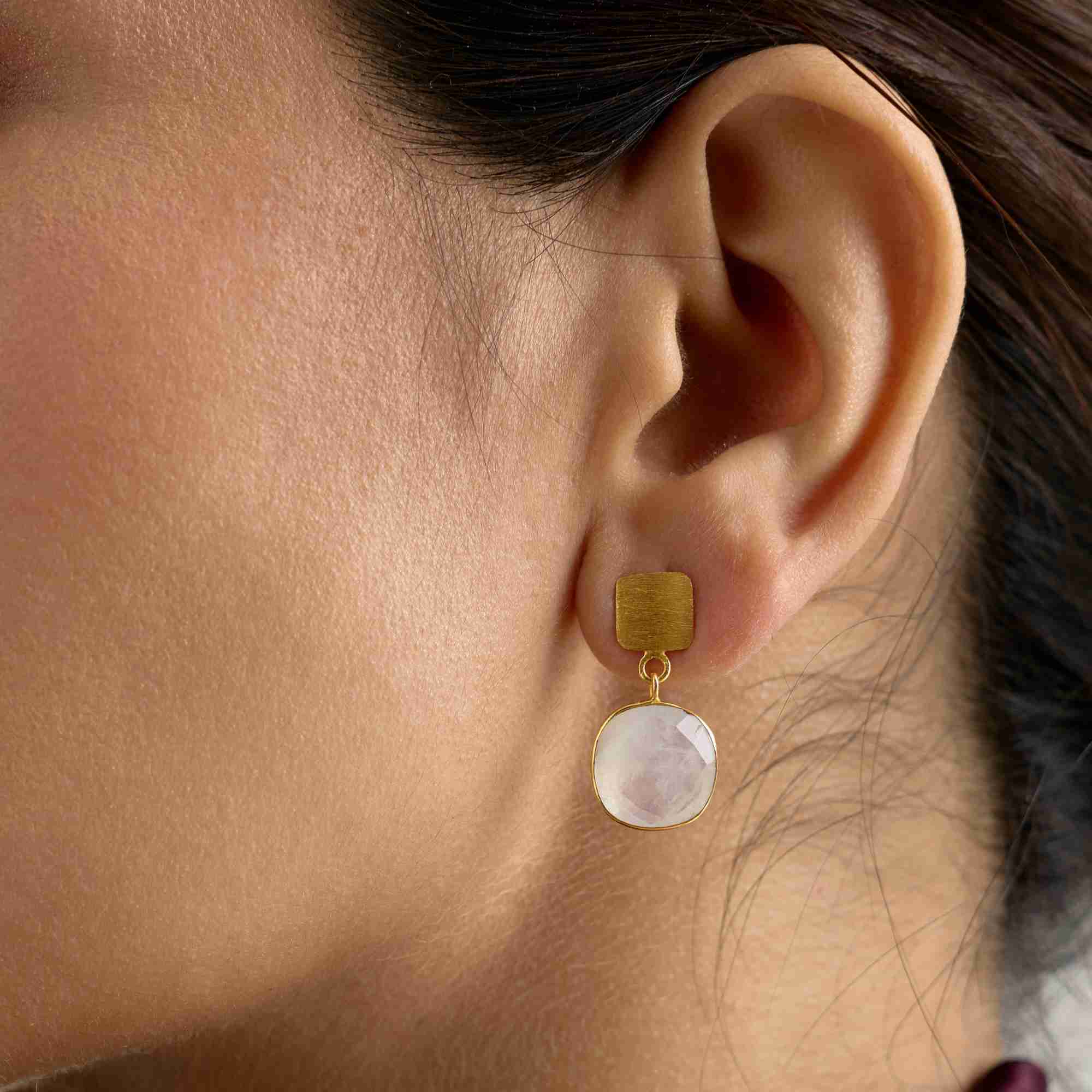 Lifestyle portrait picture named Geo Glam displaying the Cushion-Cut Rainbow Moonstone Drop Earrings Gold. A woman's profile is shown, focusing on her ear and jawline, where a pair of handcrafted 22K gold vermeil drop earrings are worn. The earrings feature a square, cushion-cut translucent Rainbow Moonstone with an ethereal blue and violet flash, suspended from a textured gold post. The model's skin is clean, and her dark hair is partially visible against a subtle background of white textiles, with soft, n