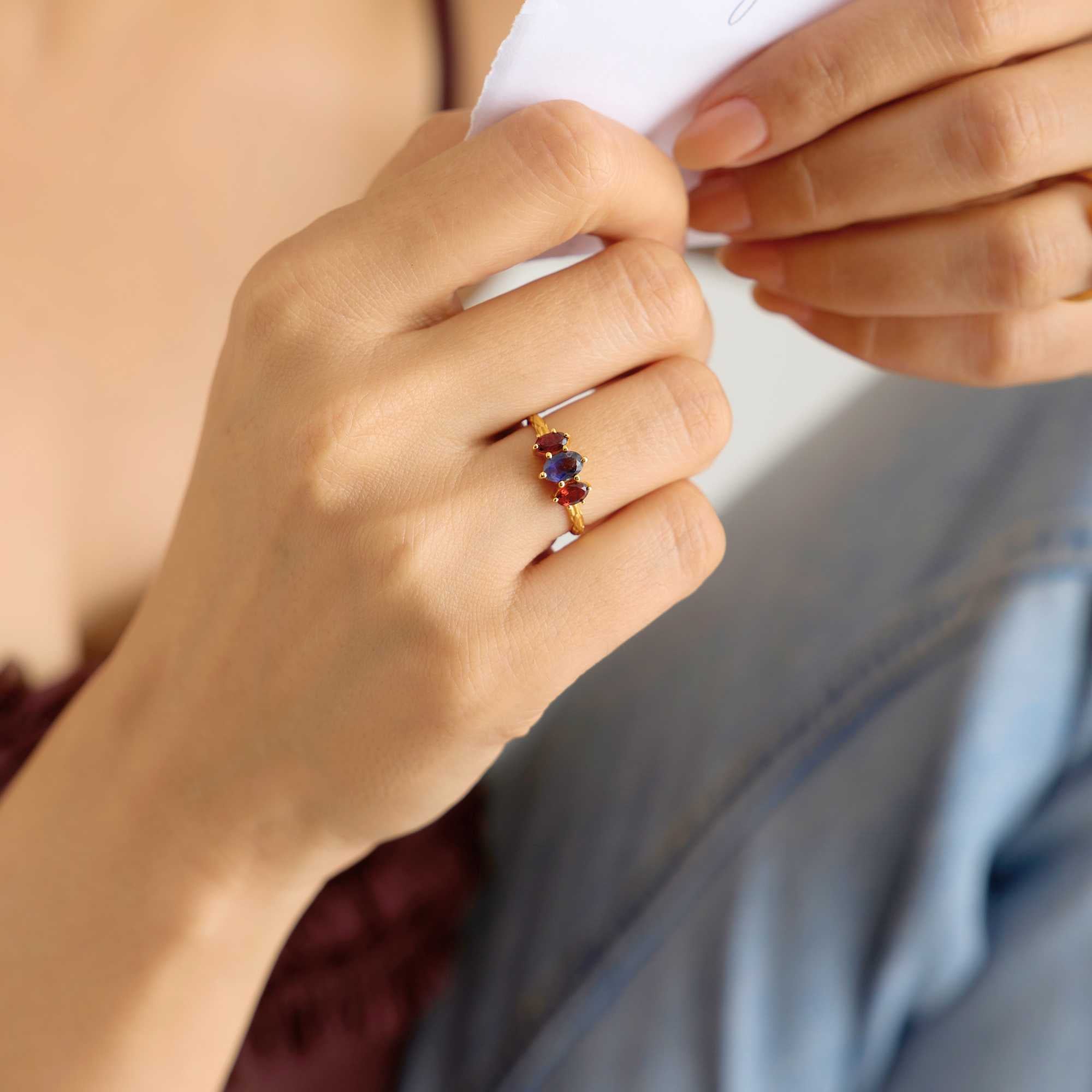 Lifestyle portrait focusing on a model's hand as she holds a piece of white paper, showcasing the Kaleidoscope Iolite and Garnet Three-Stone Marquise Ring Gold on her ring finger. The textured gold band features a central oval-cut blue iolite flanked by two deep red marquise-cut garnets.