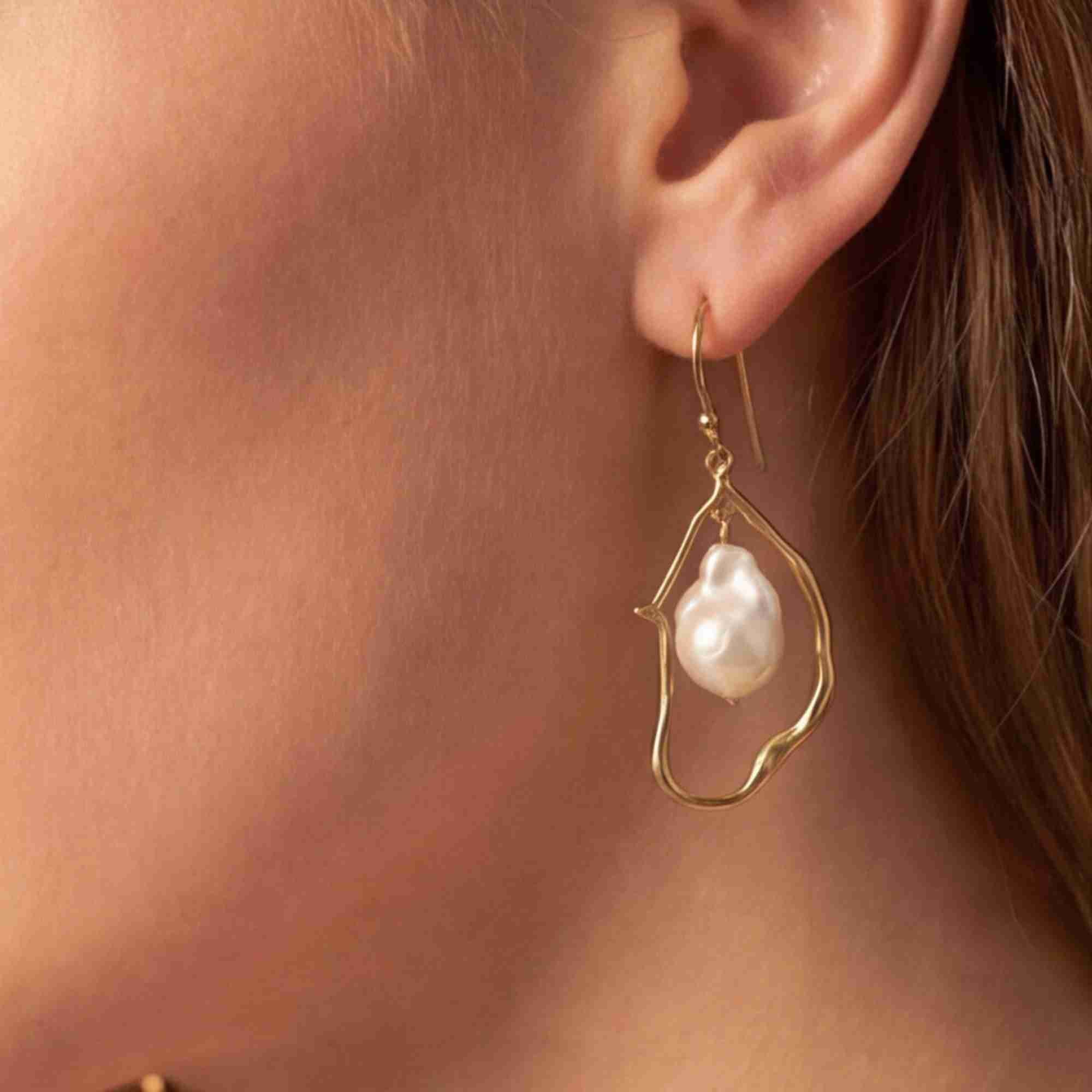 Close-up lifestyle view of a model's ear wearing the PEARLS GALORE Baroque Pearl Drop Statement Earrings Gold, referencing file name Pearl_Galore_16.jpg. The shot highlights a large, glossy white baroque pearl suspended inside a fluid, abstract molten gold wire loop frame.