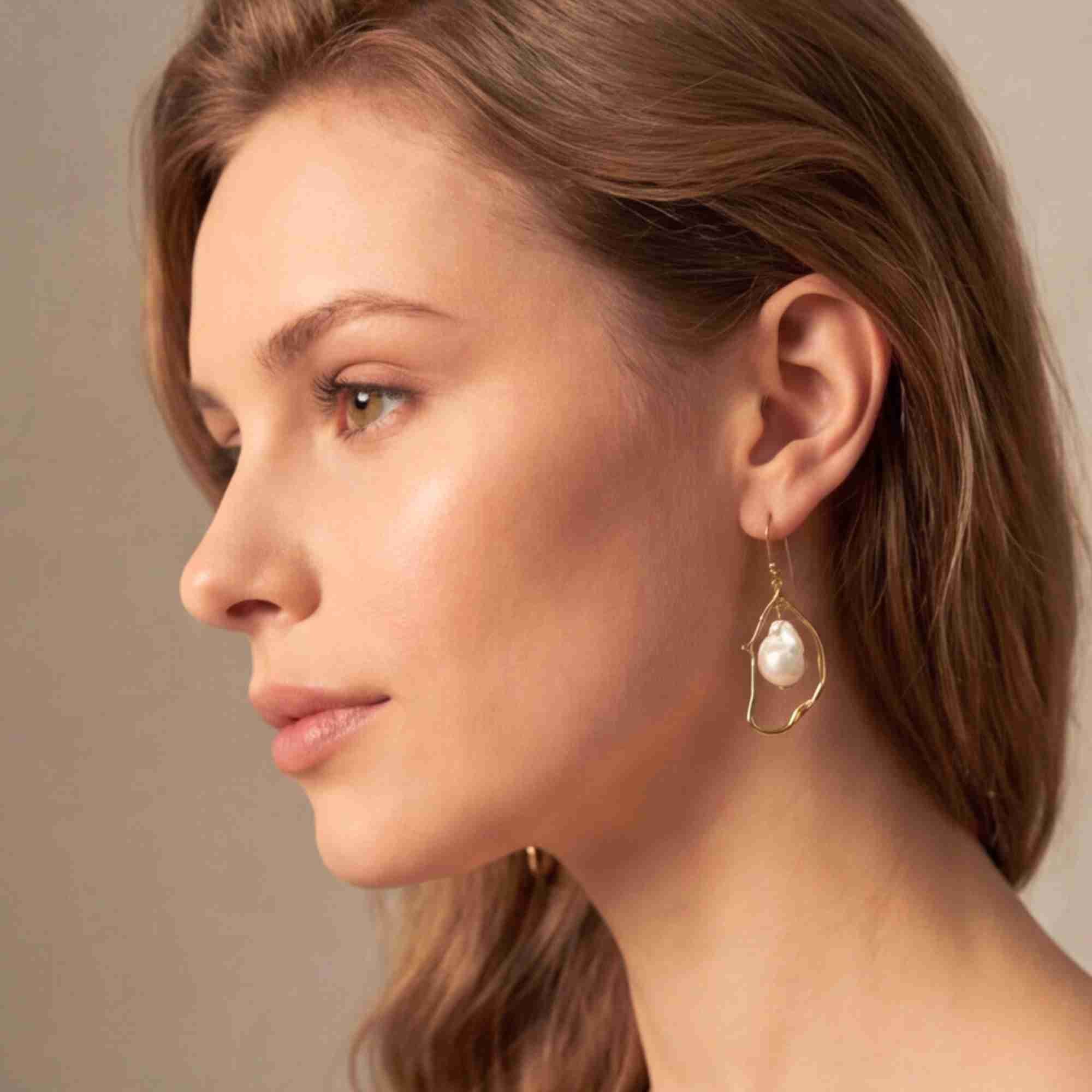 Side profile portrait of a fair-skinned model showcasing the PEARLS GALORE Baroque Pearl Drop Statement Earrings Gold, referencing file name Pearl_Galore_20.jpg. The soft studio lighting emphasizes her profile and the artistic design of the abstract gold wire drop earring with its central baroque pearl.