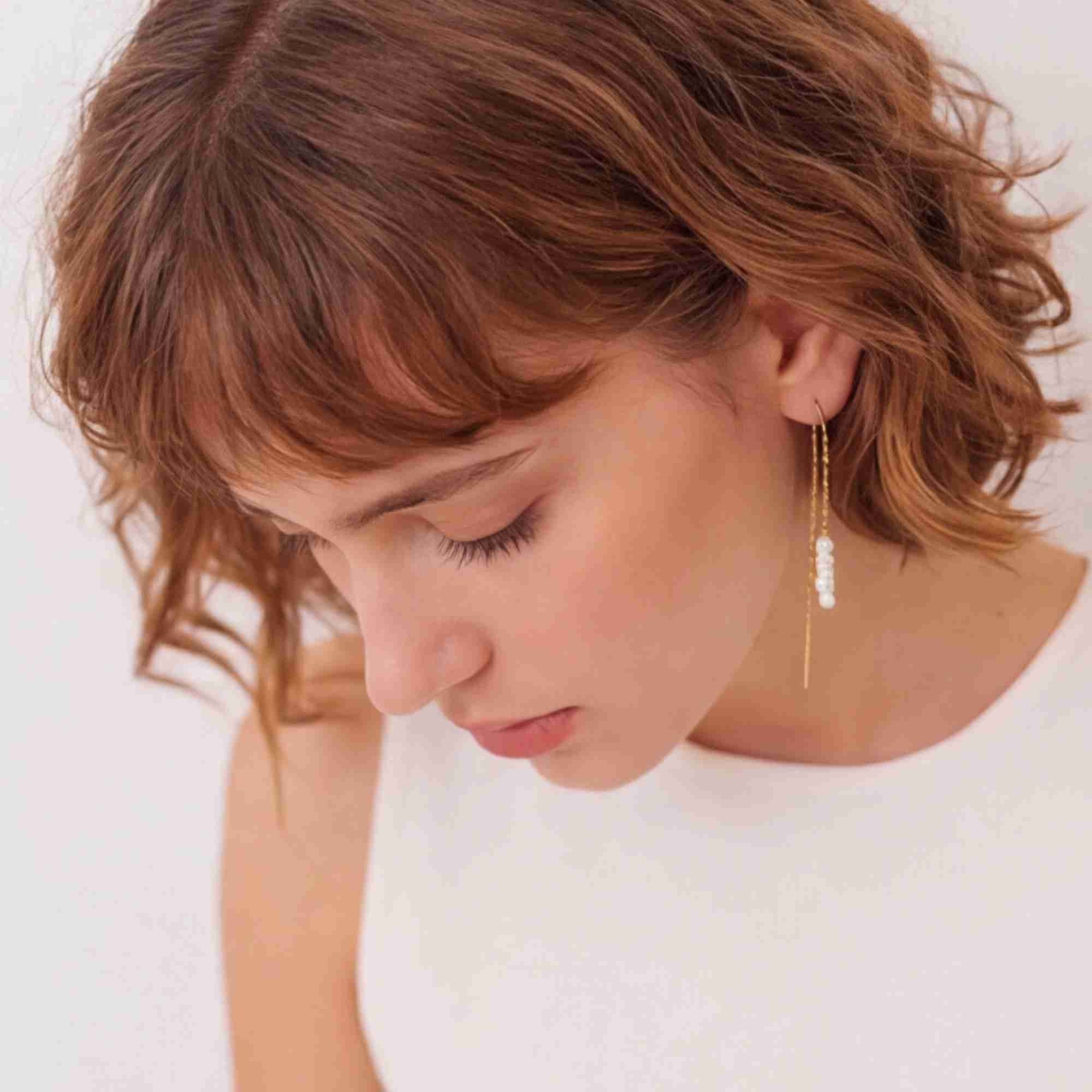 Three-quarter lifestyle view of a model looking down, highlighting the PEARLS GALORE Stacked Seed Pearl Chain Threader Earrings Gold, referencing file name Pearl_Galore_14.jpg. The soft overhead lighting emphasizes her right profile and the neat arrangement of stacked white pearls hanging against the fine gold link chain.