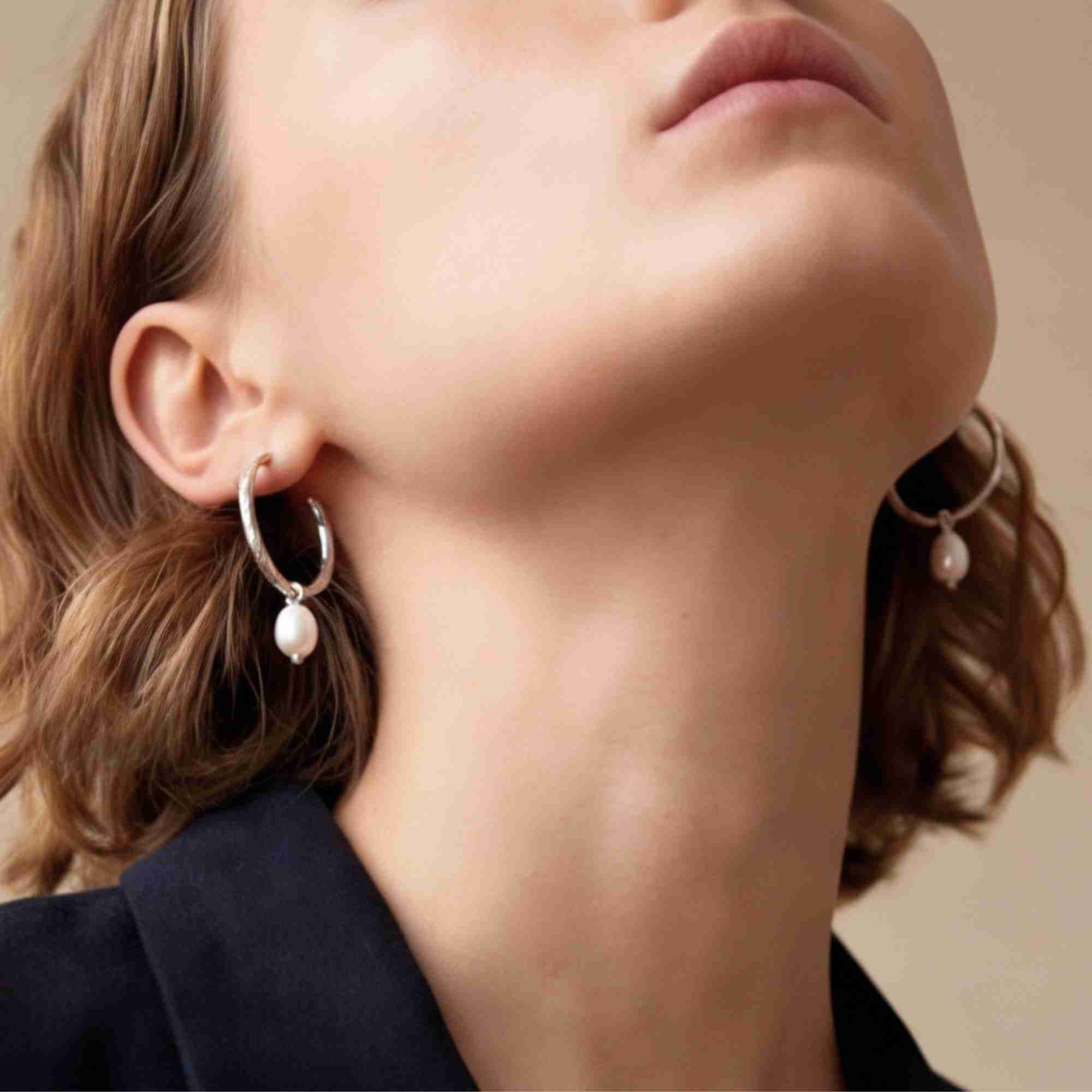 Low-angle lifestyle portrait of a model tilting her head back, displaying the PEARLS GALORE Teardrop Hoop Earrings Silver on both ears, referencing file name Pearl_Galore_9.jpg. Dressed in a dark navy blue shirt, the pose accentuates the graceful drop of the teardrop pearls suspended from the textured silver hoops.