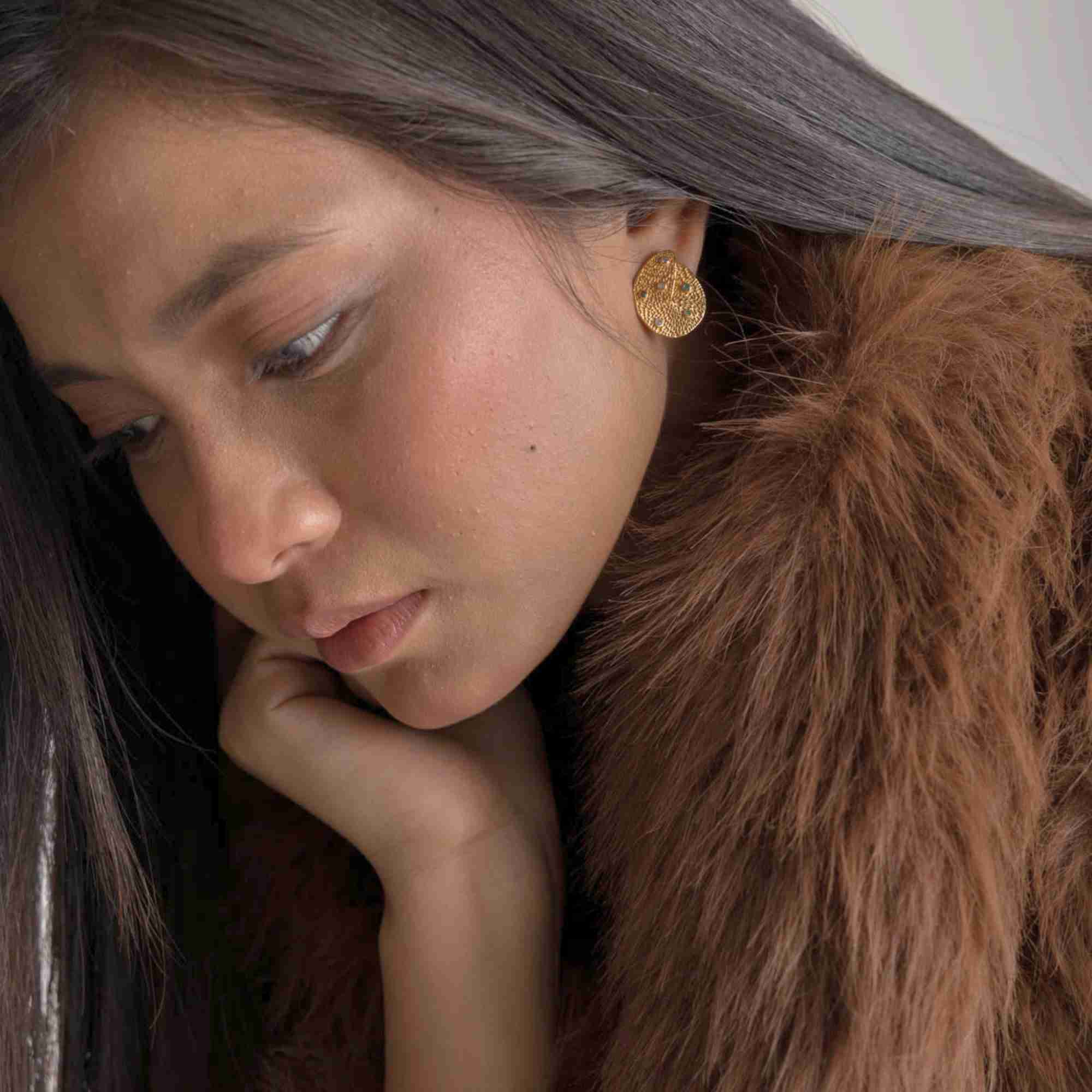 Softly lit lifestyle studio view of a woman wearing the Ribbons and Lace Galaxy Multi-Gemstone Stud Earrings Gold, referencing file name Ribbon_Lace_17.jpg. Looking downward with her hand under her chin, the pose accentuates the elegant, luxury aesthetic of the textured gold disc earring paired with dark, flowing hair.