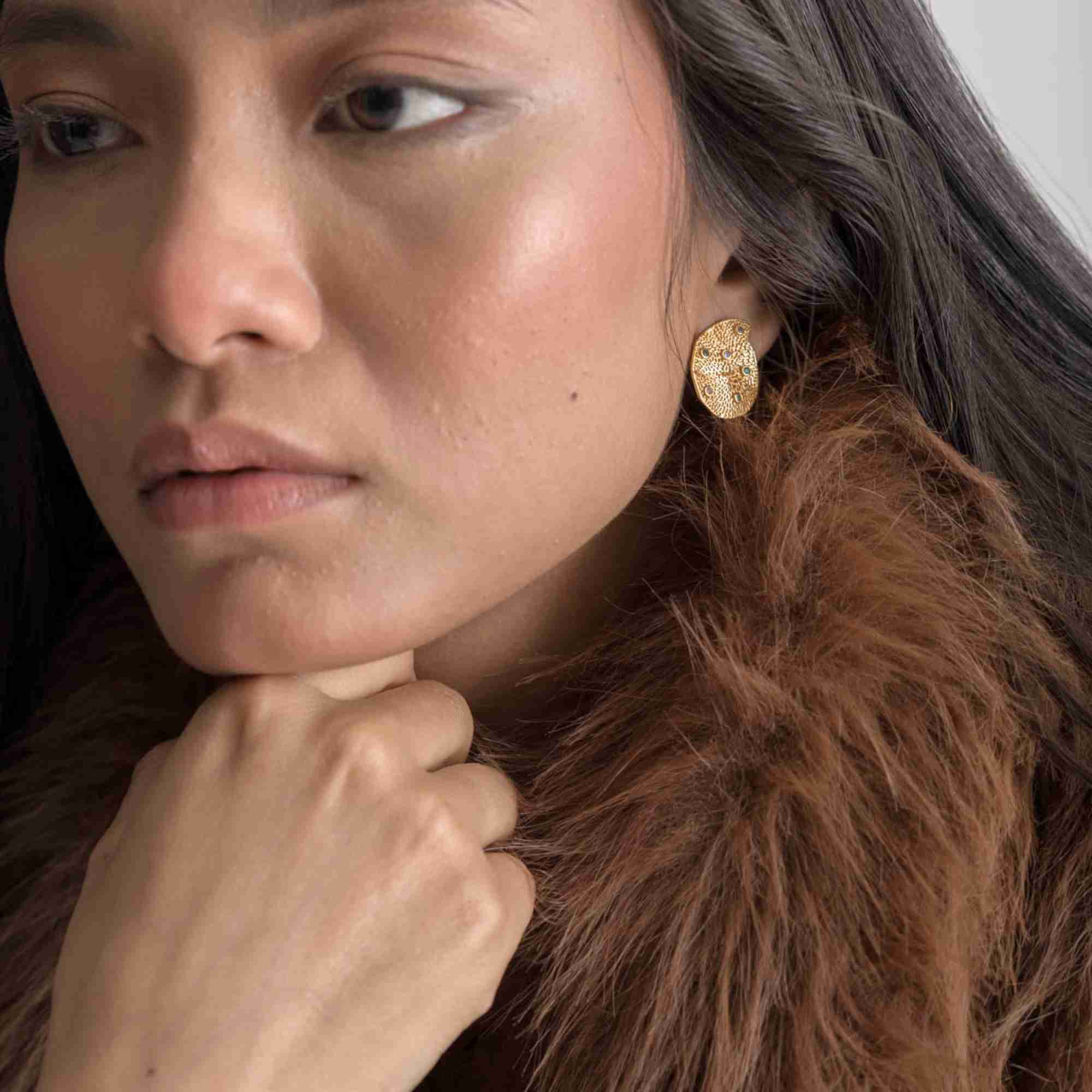 Angled lifestyle portrait of a woman modeling the Ribbons and Lace Galaxy Multi-Gemstone Stud Earrings Gold, referencing file name Ribbon_Lace_22.jpg. The shot highlights her left profile, emphasizing the textured surface detailing and elegant, flat fit of the gold abstract cluster stud earring against her earlobe.
