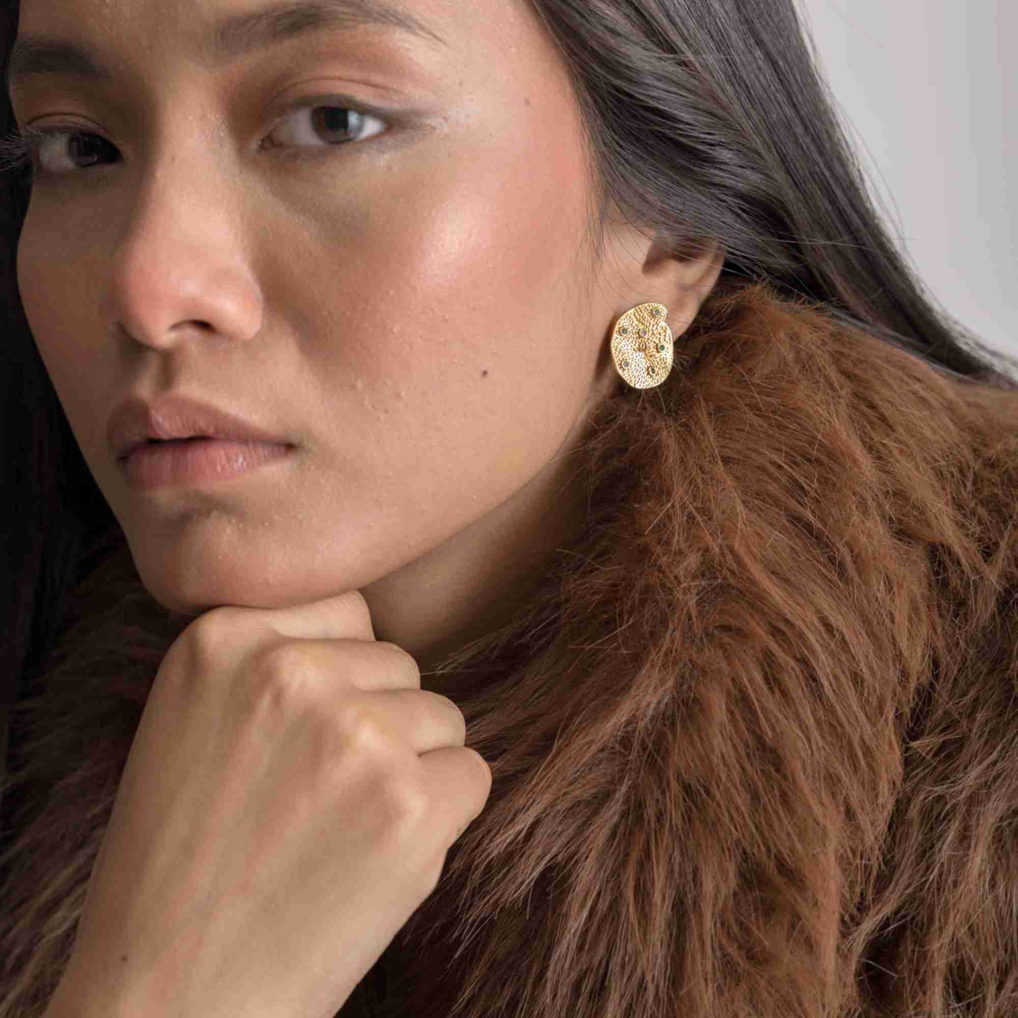 Close-up lifestyle model shot showcasing the Ribbons and Lace Galaxy Multi-Gemstone Stud Earrings Gold, referencing file name Ribbon_Lace_23.jpg. The model rests her chin on her hand, drawing focus to the intricate, lace-textured yellow gold finish and embedded galaxy-inspired colorful gems of the statement stud earring.