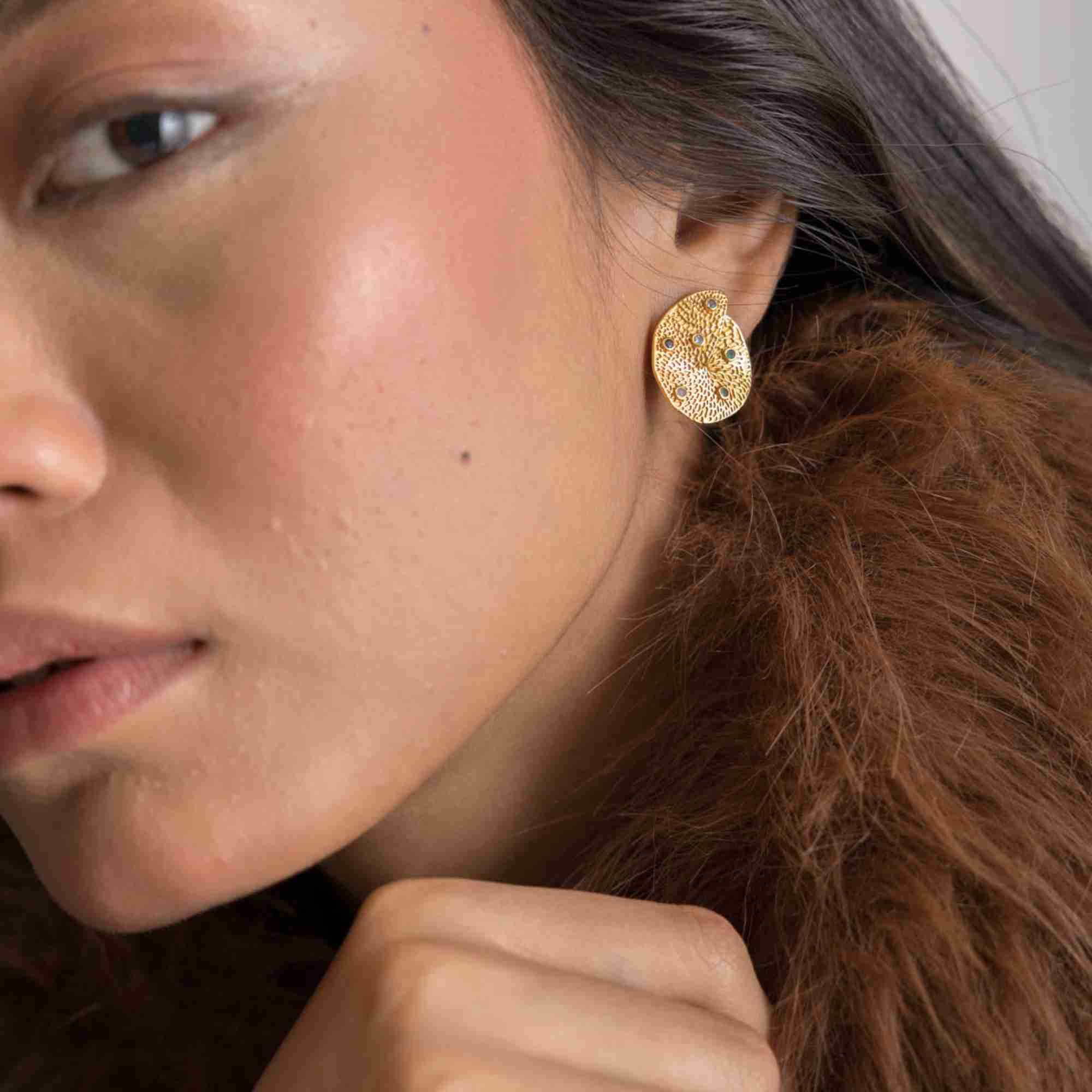 Detailed side-profile lifestyle view of the Ribbons and Lace Galaxy Multi-Gemstone Stud Earrings Gold, referencing file name Ribbon_Lace_20.jpg. The close-up captures the shimmering, fine-textured pattern and tiny inset multi-colored gemstones scattered across the curved organic gold disc.