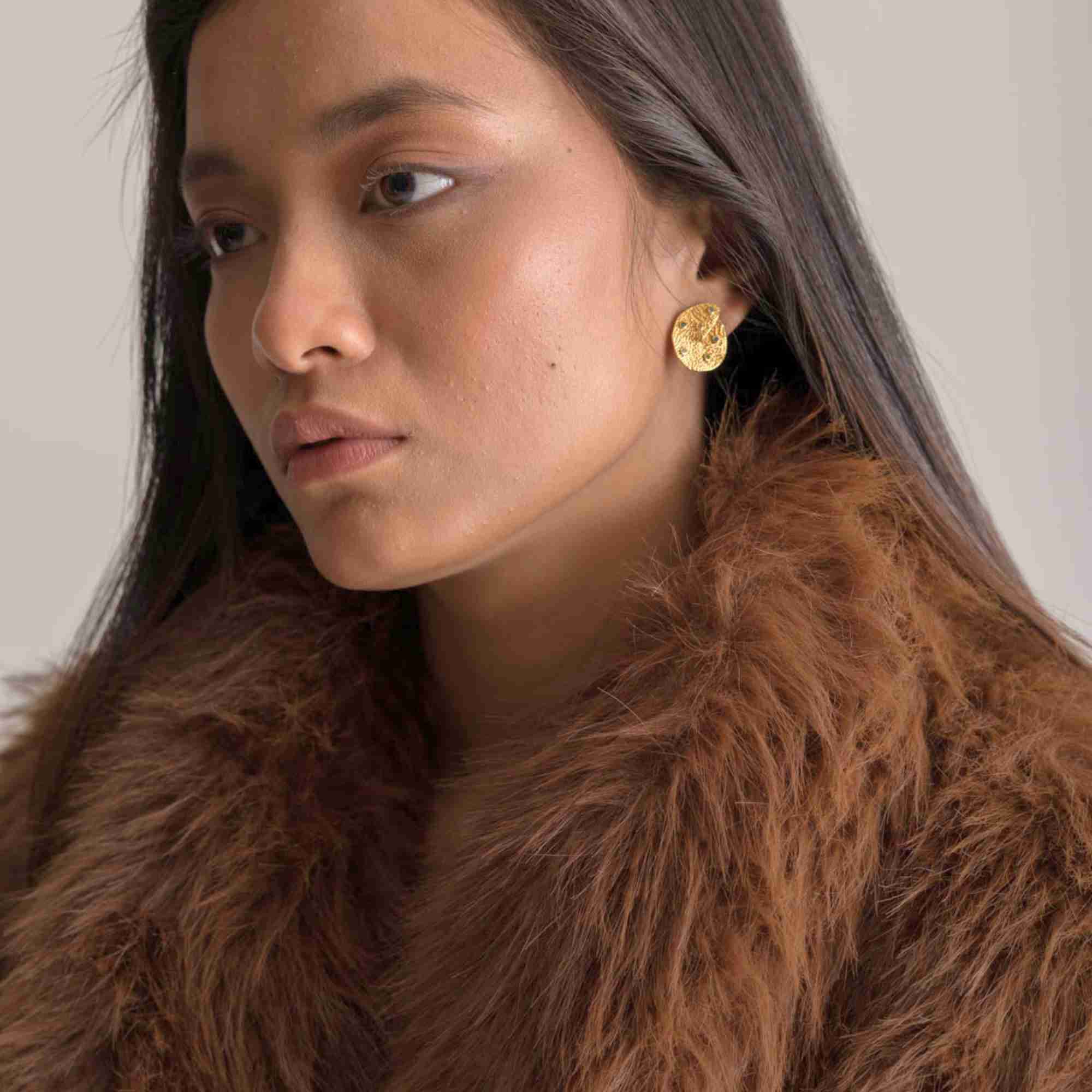 Three-quarter lifestyle portrait of a model accessorized with the Ribbons and Lace Galaxy Multi-Gemstone Stud Earrings Gold, referencing file name Ribbon_Lace_16.jpg. The composition focuses on her facial profile and the upper torso, highlighting how the statement gold cosmic stud earring acts as a striking piece against the rich texture of her brown fur outfit.