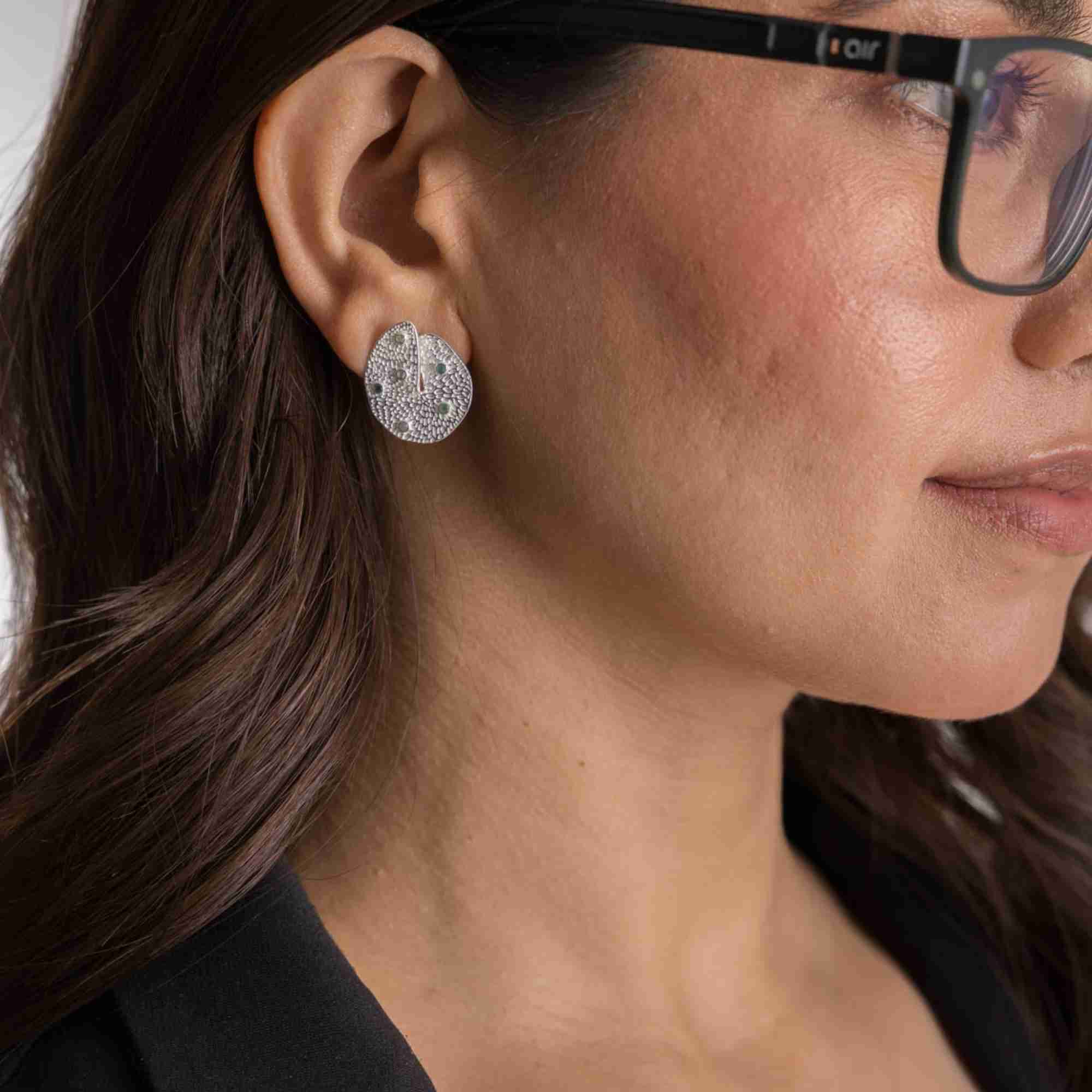 Close-up lifestyle view of a woman wearing the Ribbons and Lace Galaxy Multi-Gemstone Stud Earrings Silver, referencing file name Ribbon_Lace_16_6ab60015-ef64-4b35-8757-710507d1cacd.jpg. The shot displays her right ear featuring an organic, textured oval silver disc stud studded with tiny multi-colored gemstones. She is wearing black-rimmed glasses and a black blazer against a light background.