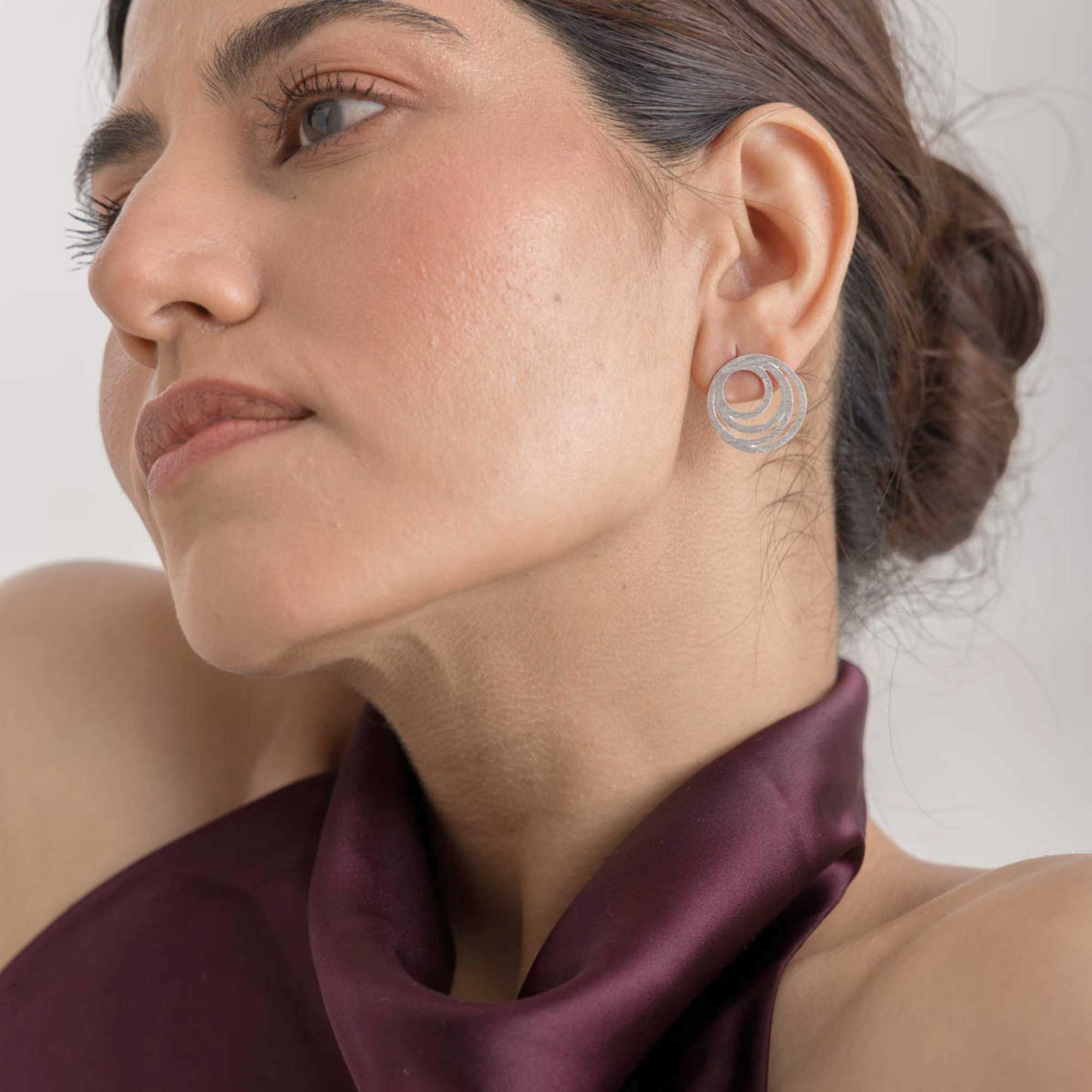 Front-facing lifestyle model view showcasing the Shimmering Disks Intermingled Circles Stud Earrings Silver, referencing file name Shimmering Disks (71).jpg. The model tilts her head slightly to the side, drawing attention to the geometric nested-circle design of the high-shine 92.5 sterling silver earring resting comfortably against her earlobe.