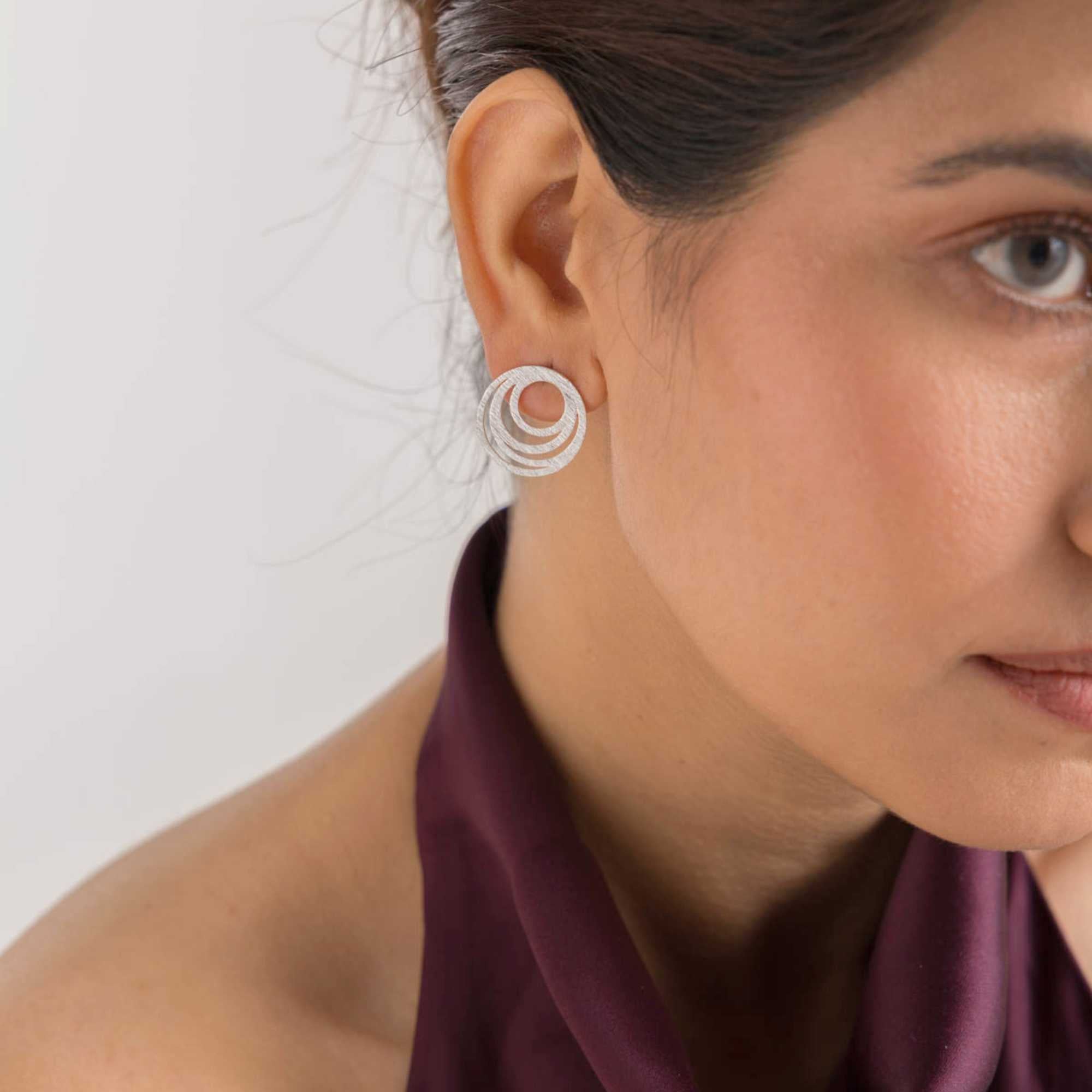 Lifestyle close-up view of the Shimmering Disks Intermingled Circles Stud Earrings Silver on a model, referencing file name 28_94f5e20a-b3b6-4c3a-93db-cf5abfa41caf.jpg. The shot highlights a woman's right ear wearing a round sterling silver stud earring adorned with multiple textured, nested concentric circles. She is styled in a deep plum halter neck top against a clean, white studio background.