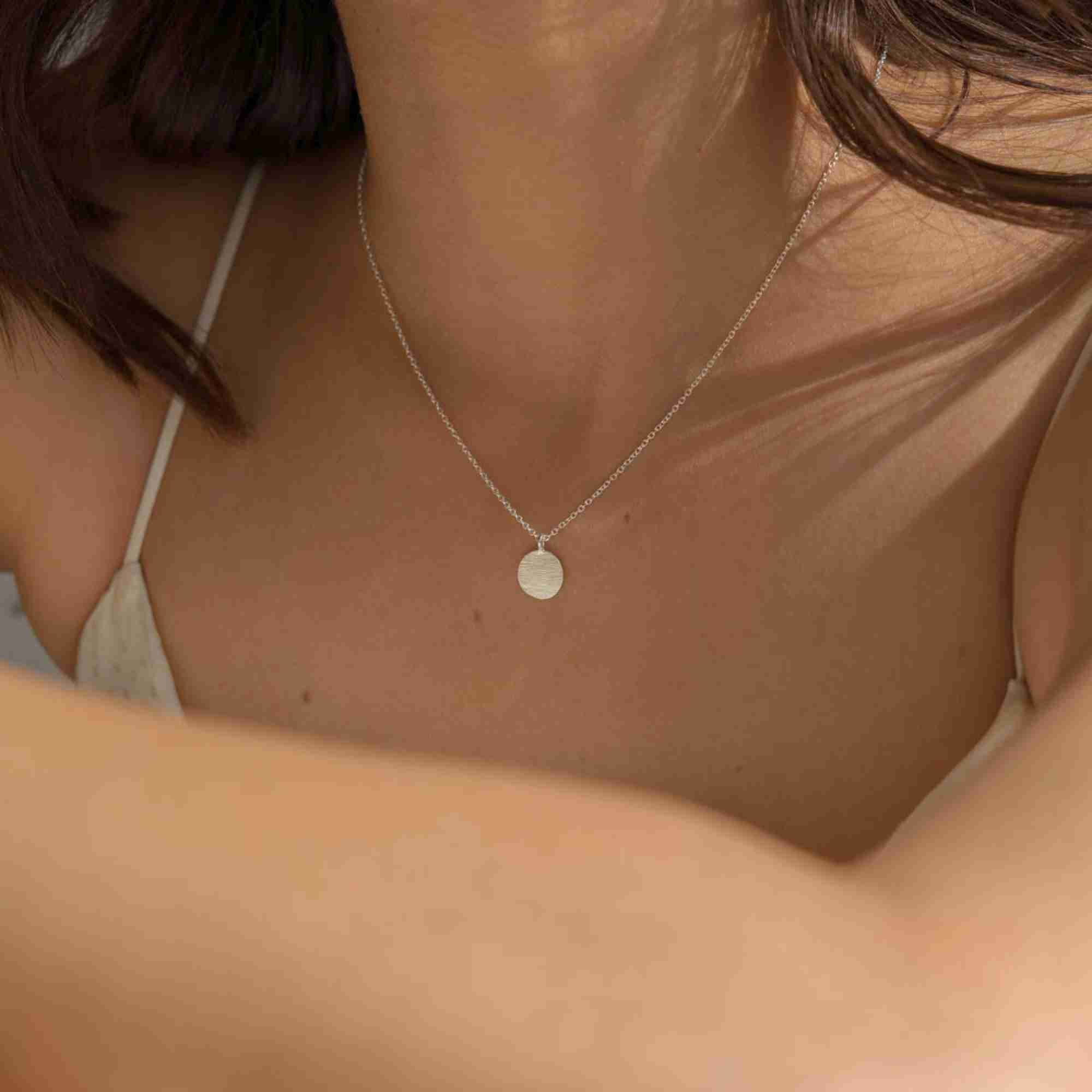 Candid lifestyle model view of the Shimmering Disks Minimalist Coin Pendant Necklace Silver, referencing file name 129.jpg. The image captures the upper torso of a woman in a cream dress with her arm subtly in the foreground, showcasing how the small, sleek silver disk disc pendant acts as a delicate daily accent piece.