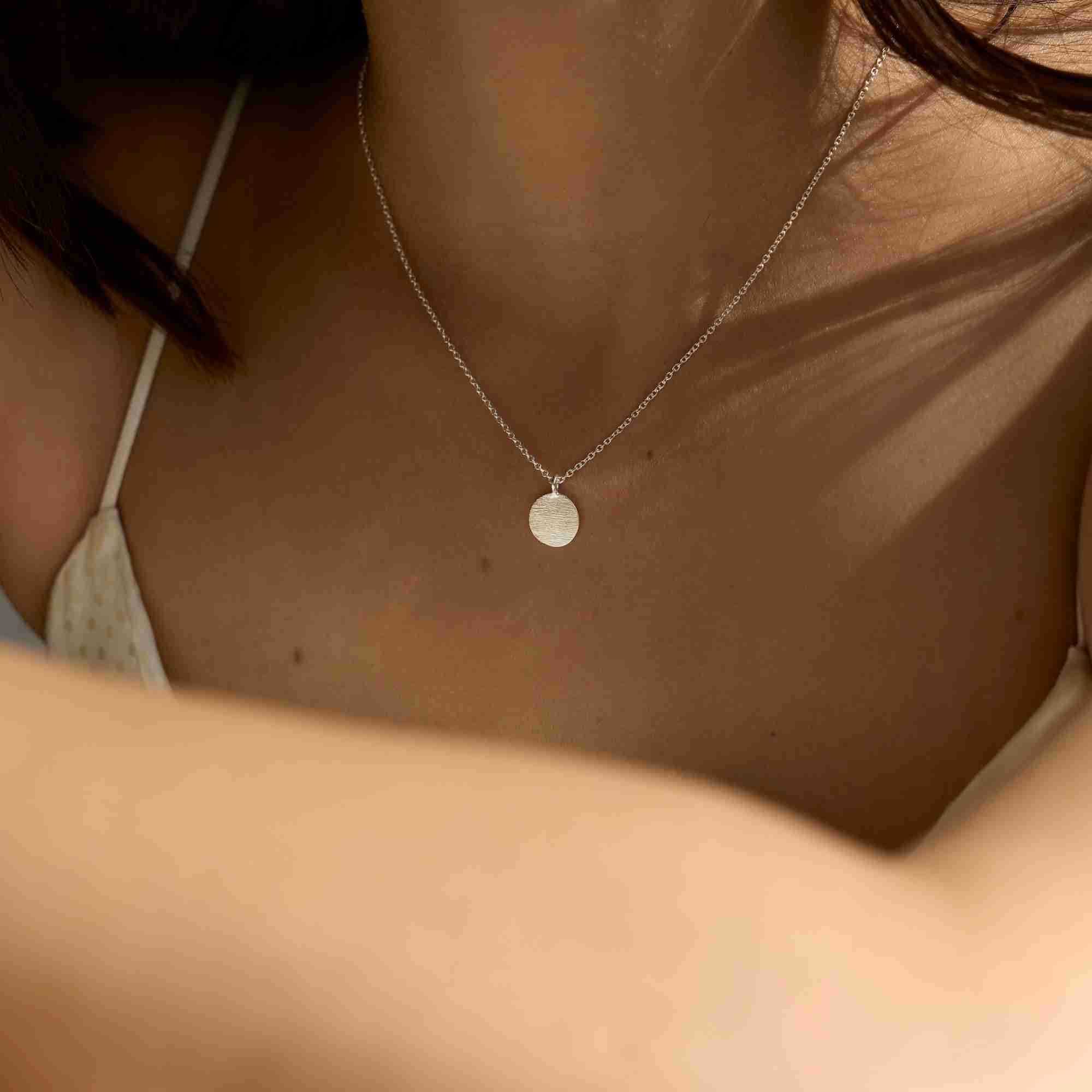 Cinematic lifestyle view of the Shimmering Disks Minimalist Coin Pendant Necklace Silver, referencing file name Shimmering_Disks_60.jpg. The model poses under dramatic sunlight and shadows, emphasizing the fine details of the high-polish circular silver coin charm as it catches the light on her chest.