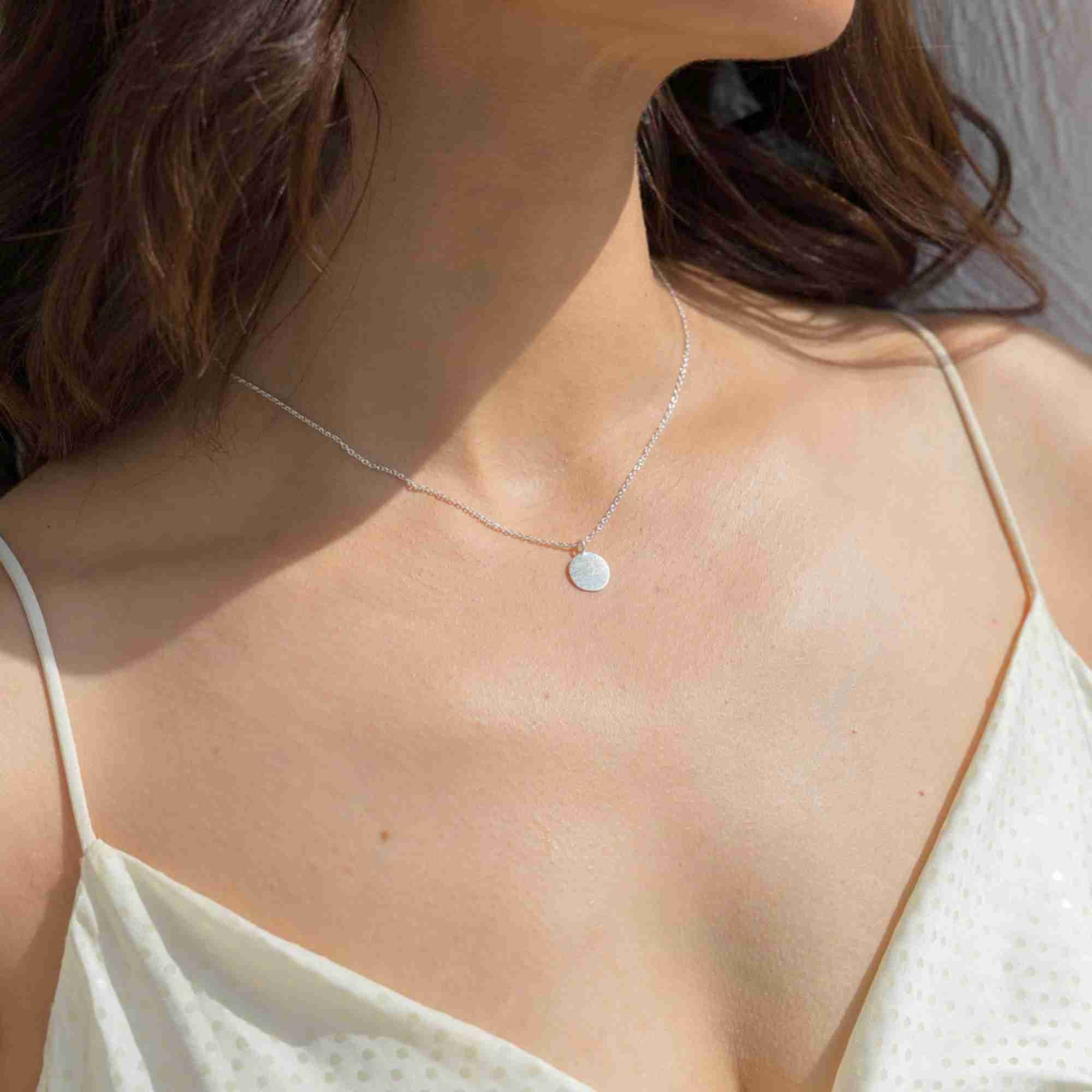 Lifestyle model view showing a three-quarter angle of a woman modeling the Shimmering Disks Minimalist Coin Pendant Necklace Silver, referencing file name 128.jpg. The minimalist piece features a simple, flat geometric circular coin tag crafted in polished recycled sterling silver suspended from a fine silver chain.