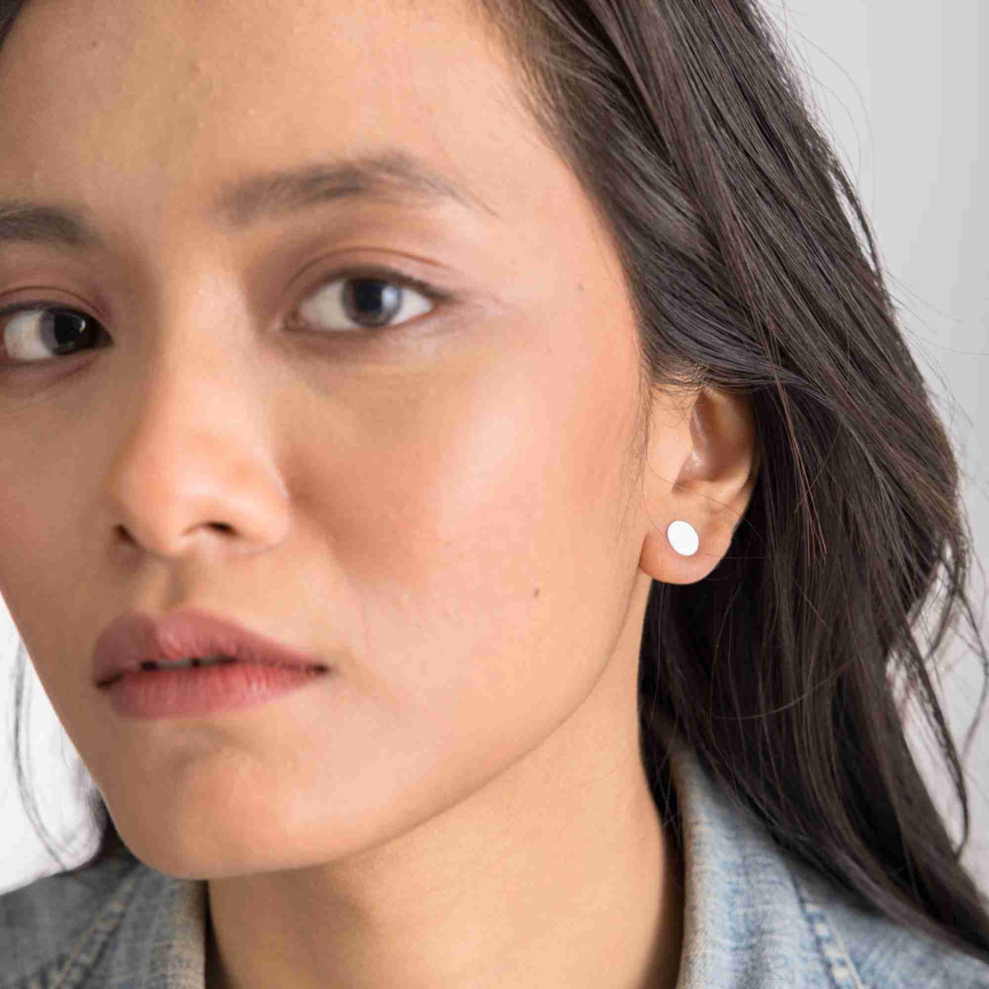 Lifestyle portrait view of a model showcasing the Shimmering Disks Minimalist Stud Earrings Silver, referencing file name Shimmering_Disks_70_1742a710-9ae7-4d6f-a4a1-ab75c1fb097a.jpg. The shot highlights a small, flat, polished circular 92.5 sterling silver disc stud earring on her left earlobe. She is styled in a casual denim jacket with her dark hair swept back.