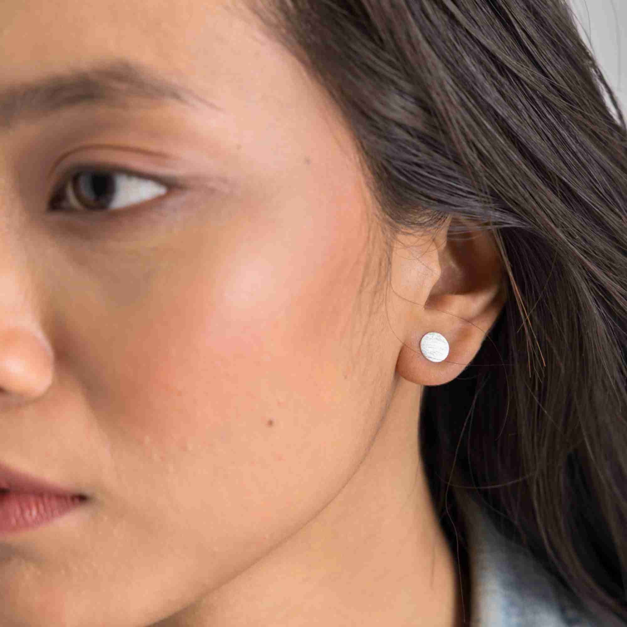 Extreme close-up lifestyle view focusing on the Shimmering Disks Minimalist Stud Earrings Silver, referencing file name Shimmering_Disks_72_c5fafa8d-7a4b-4f33-b592-be16b35a28ba.jpg. The angled detail shot captures the fine, shimmering scratch-textured finish across the flat surface of the tiny round silver stud earring.