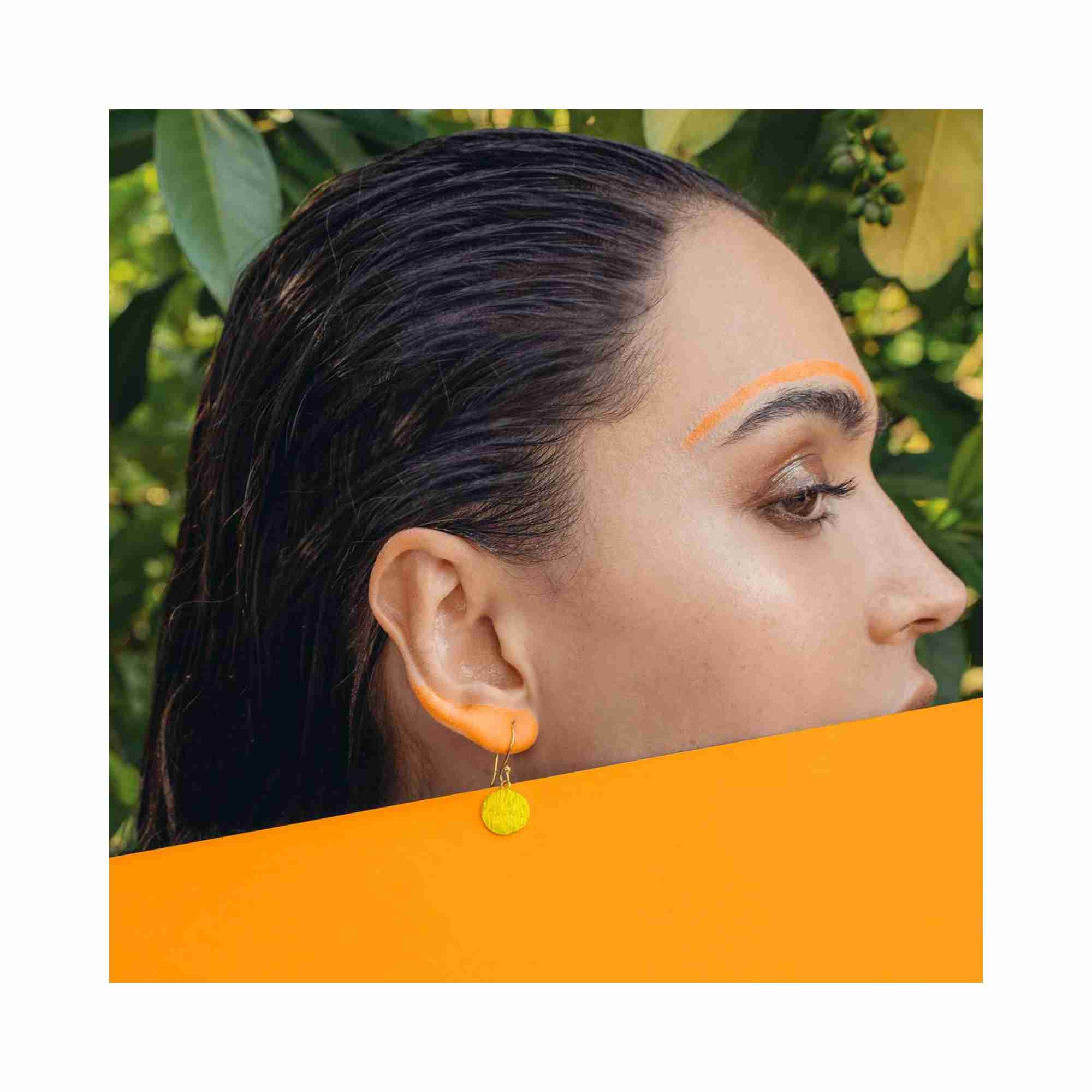 Artistic editorial photoshoot of Shimmering Disks minimalist medium gold coin drop earrings, styled on a model with sunset-orange makeup accents against a lush green nature backdrop to highlight the 22K gold vermeil shimmer.