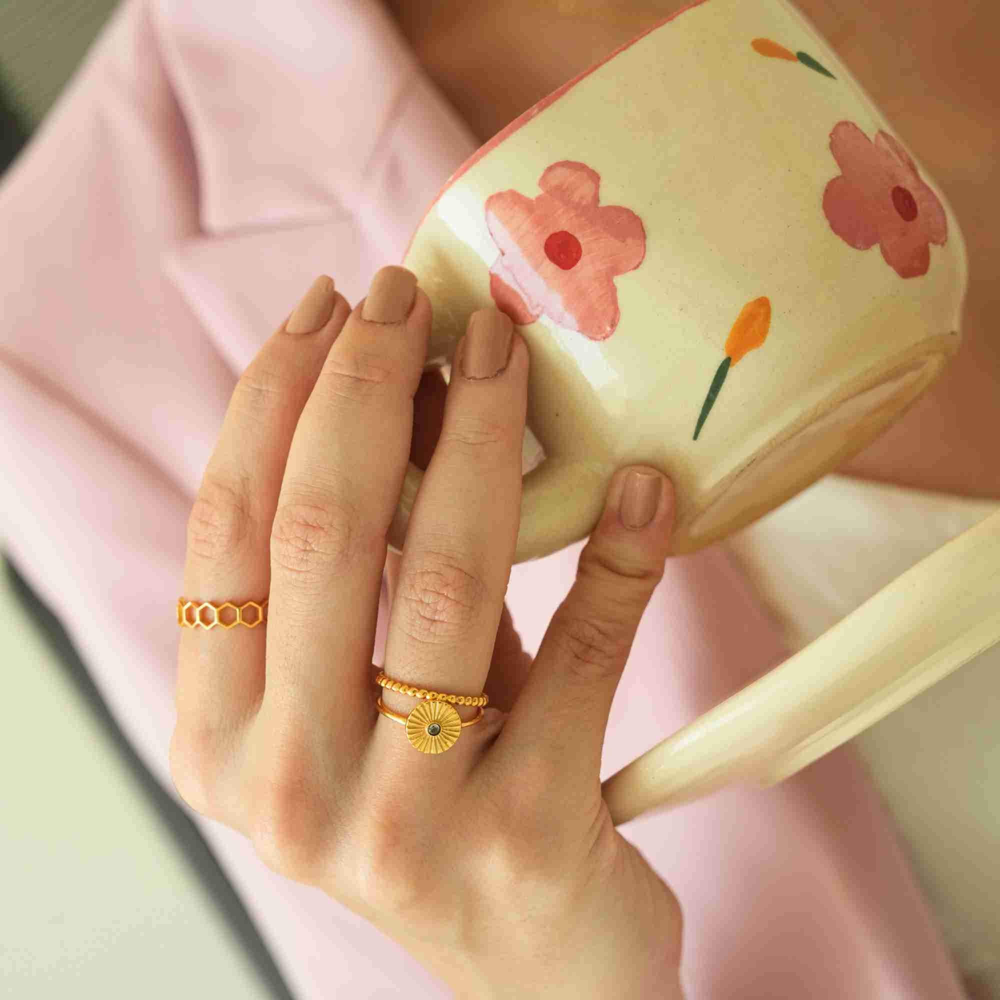 Side angle of a model's hand holding a hand-painted floral ceramic cup, highlighting the textured design of the Delicate Vintage Fluted Disc Ring in Gold by Protsaah Zurich paired with a delicate beaded gold band.