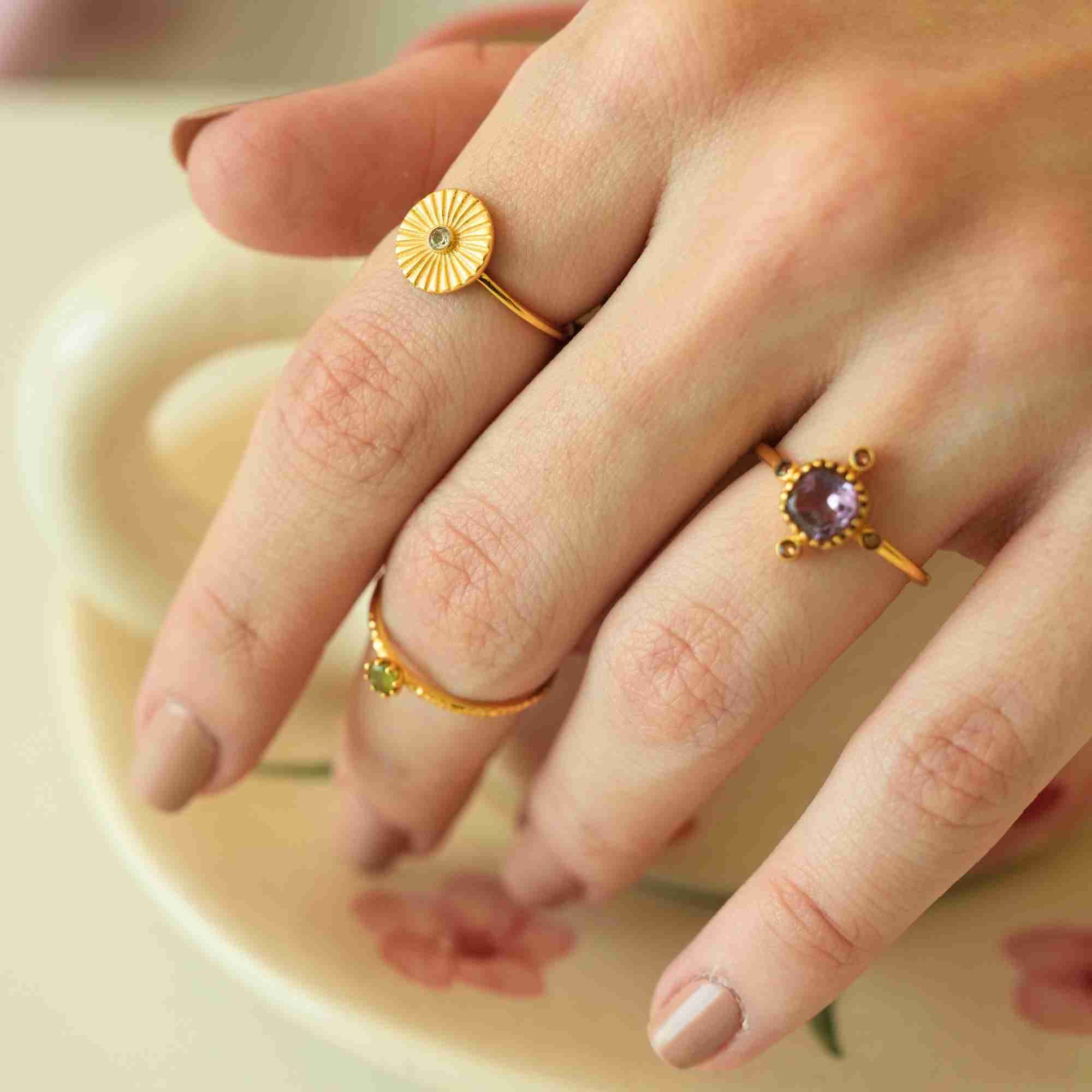 A detailed close-up of a model's hand displaying a curated ring stack, emphasizing the Delicate Vintage Fluted Disc Ring in Gold by Protsaah Zurich worn alongside statement gemstone rings over a floral ceramic plate.
