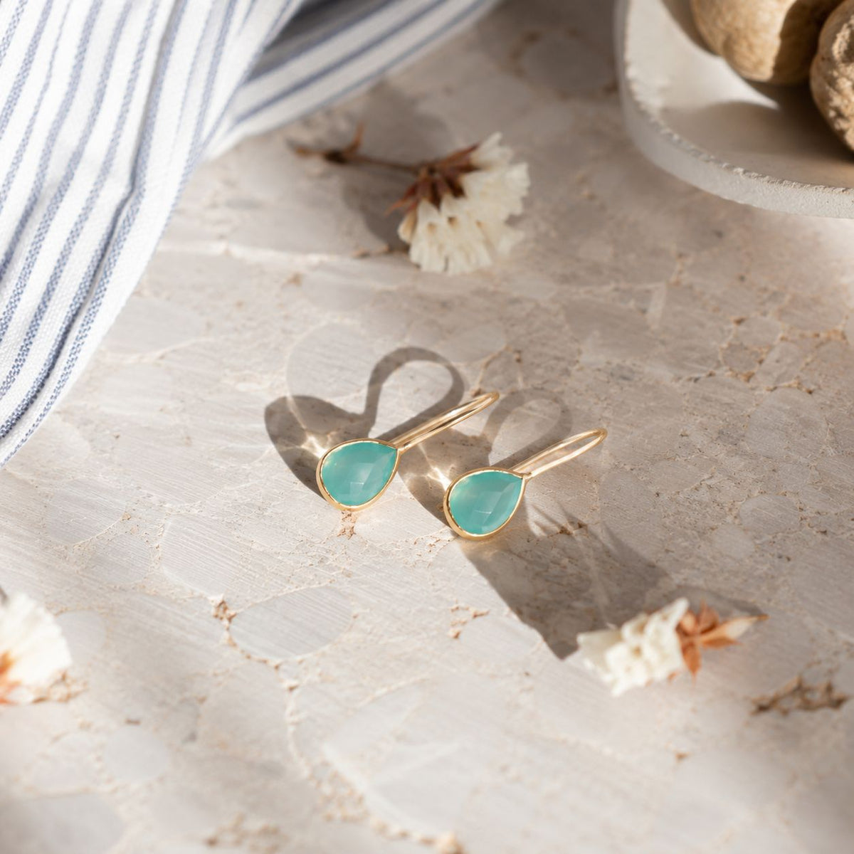 A pair of gold teardrop stud earrings with light green cabochon stones, styled on a textured beige stone surface with dried flowers and a striped fabric.