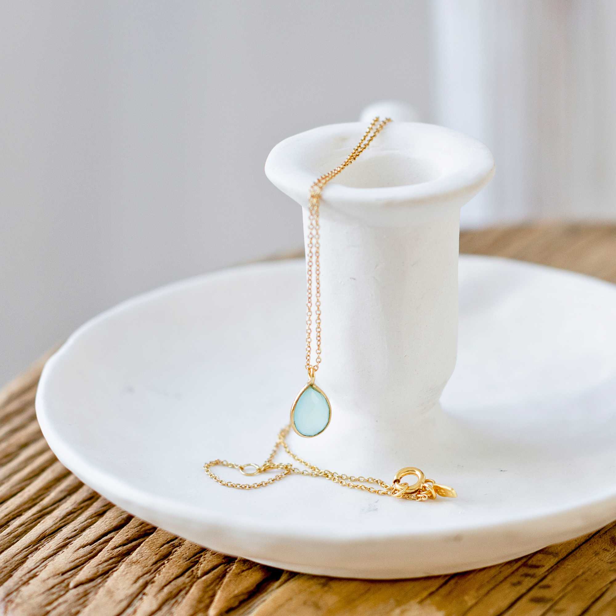 A gold pendant necklace featuring a delicate, bezel-set teardrop aqua chalcedony gemstone charm on a fine chain, shown both displayed on a white ceramic candlestick holder and worn by a model.