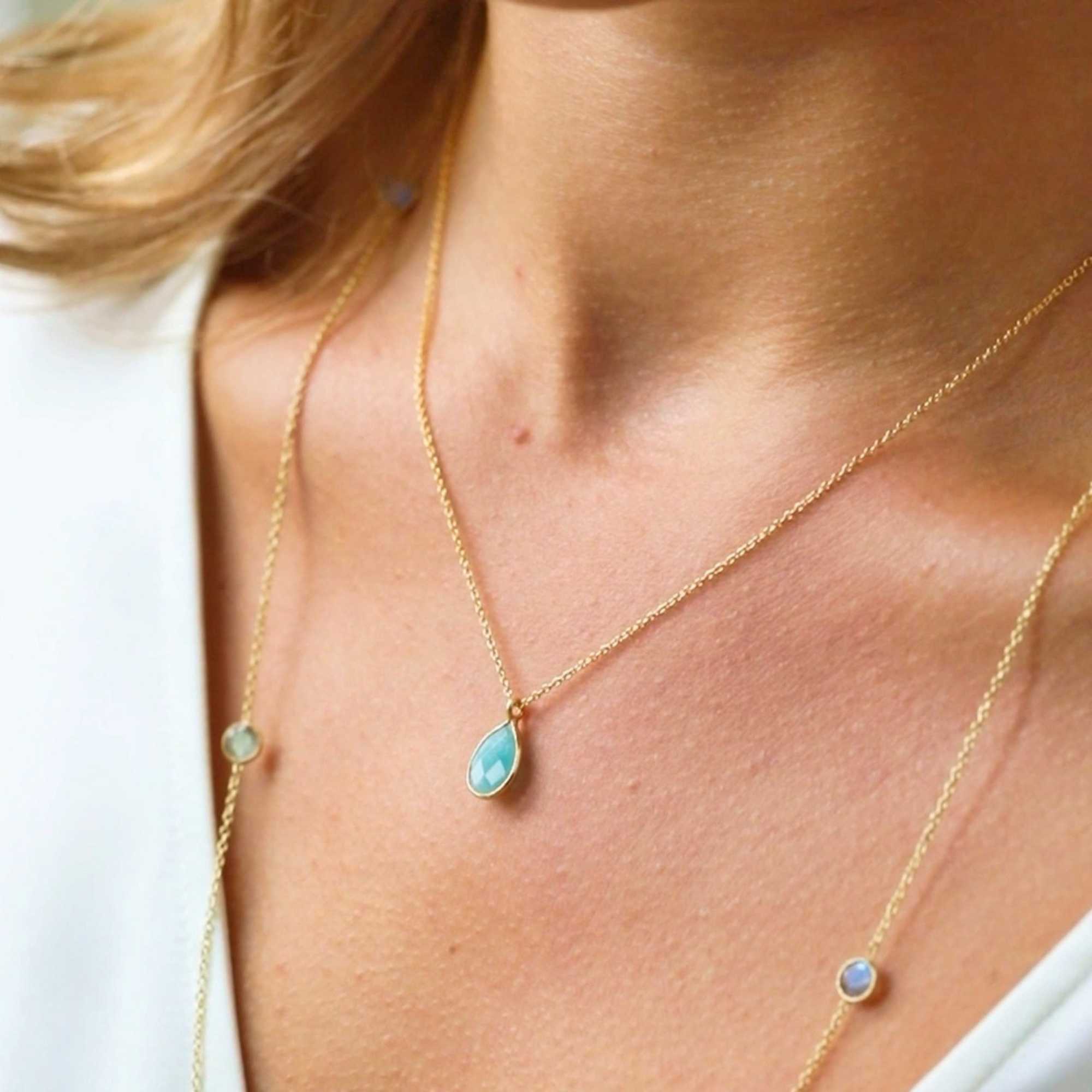 A gold pendant necklace featuring a delicate, bezel-set teardrop aqua chalcedony gemstone charm on a fine chain, shown both displayed on a white ceramic candlestick holder and worn by a model.