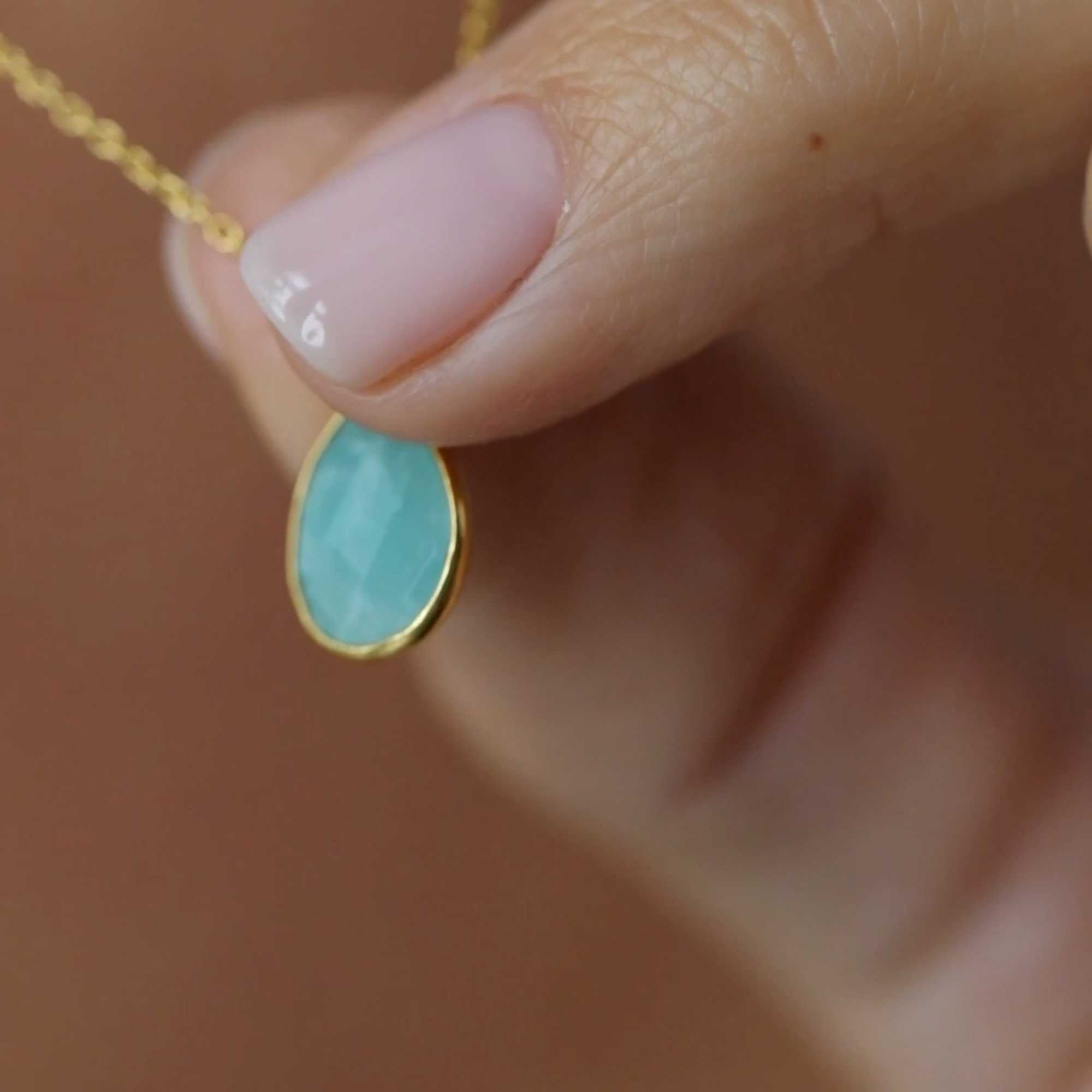 A gold pendant necklace featuring a delicate, bezel-set teardrop aqua chalcedony gemstone charm on a fine chain, shown both displayed on a white ceramic candlestick holder and worn by a model.