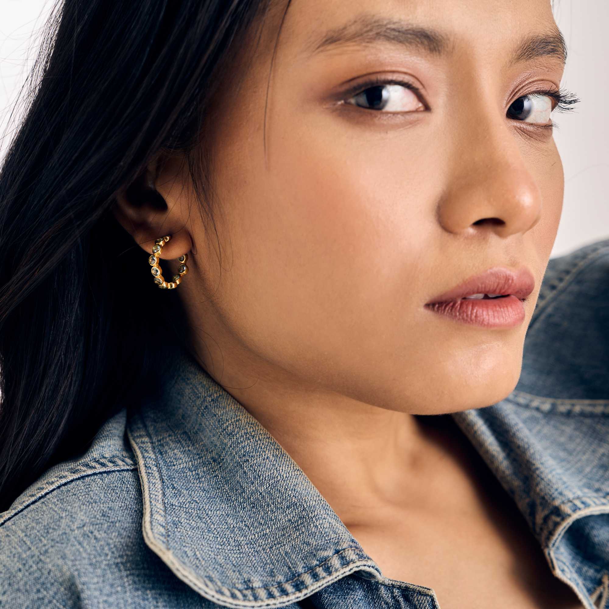 Gold hoop earrings studded with round, bezel-set blue topaz gemstones, shown worn on a model paired with a denim jacket.
