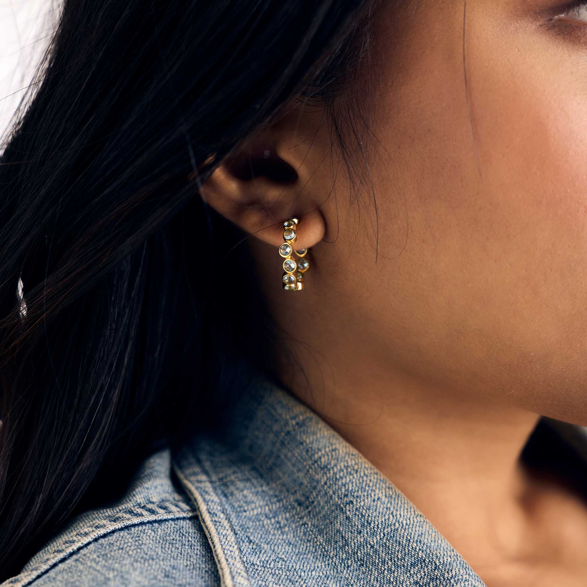 Gold hoop earrings studded with round, bezel-set blue topaz gemstones, shown worn on a model paired with a denim jacket.
