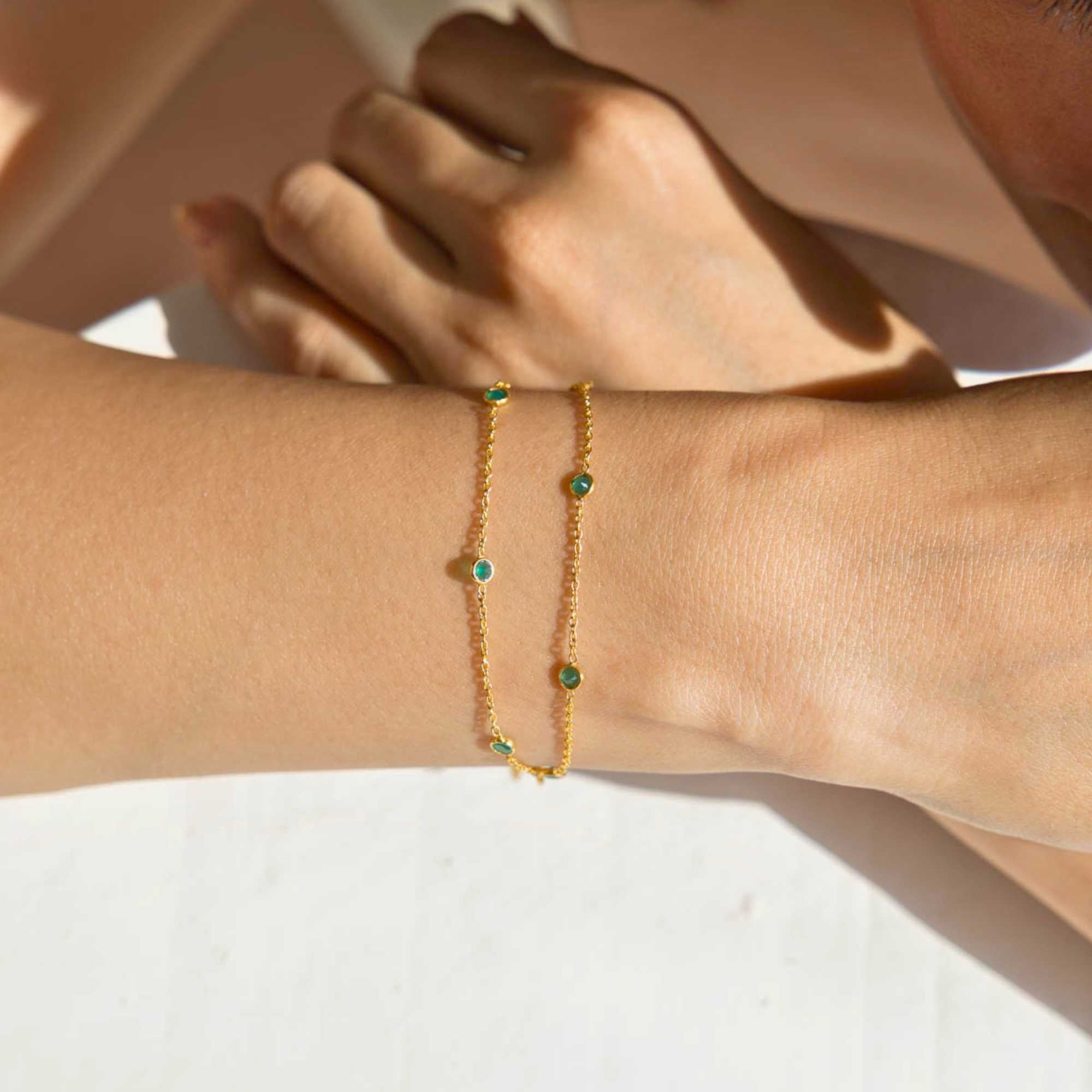 A close-up lifestyle shot of a person's wrist wearing the Dew Drops Green Onyx Studded Chain Bracelet Gold, showcasing the delicate double gold chains and bezel-set green onyx gemstones under warm, natural sunlight.