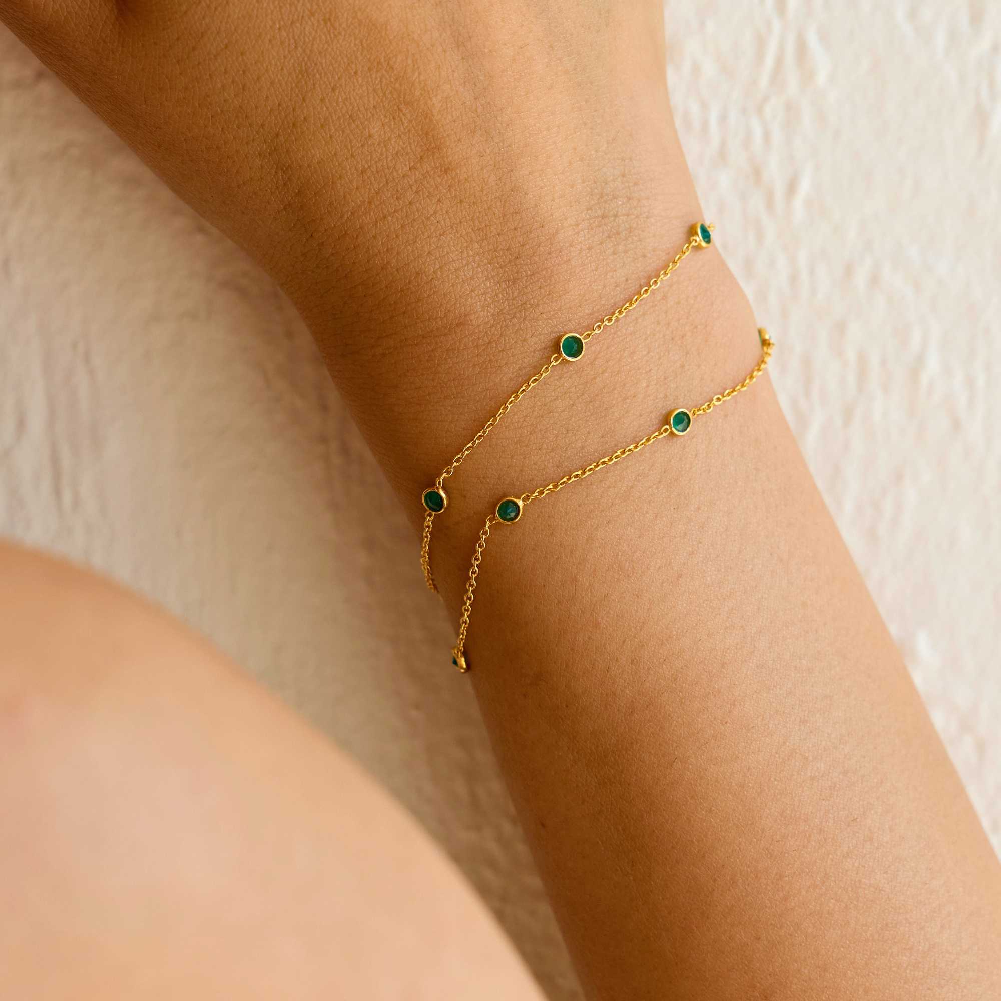 A close-up lifestyle shot of a person's wrist wearing the Dew Drops Green Onyx Studded Chain Bracelet Gold, showcasing the delicate double gold chains and bezel-set green onyx gemstones under warm, natural sunlight.