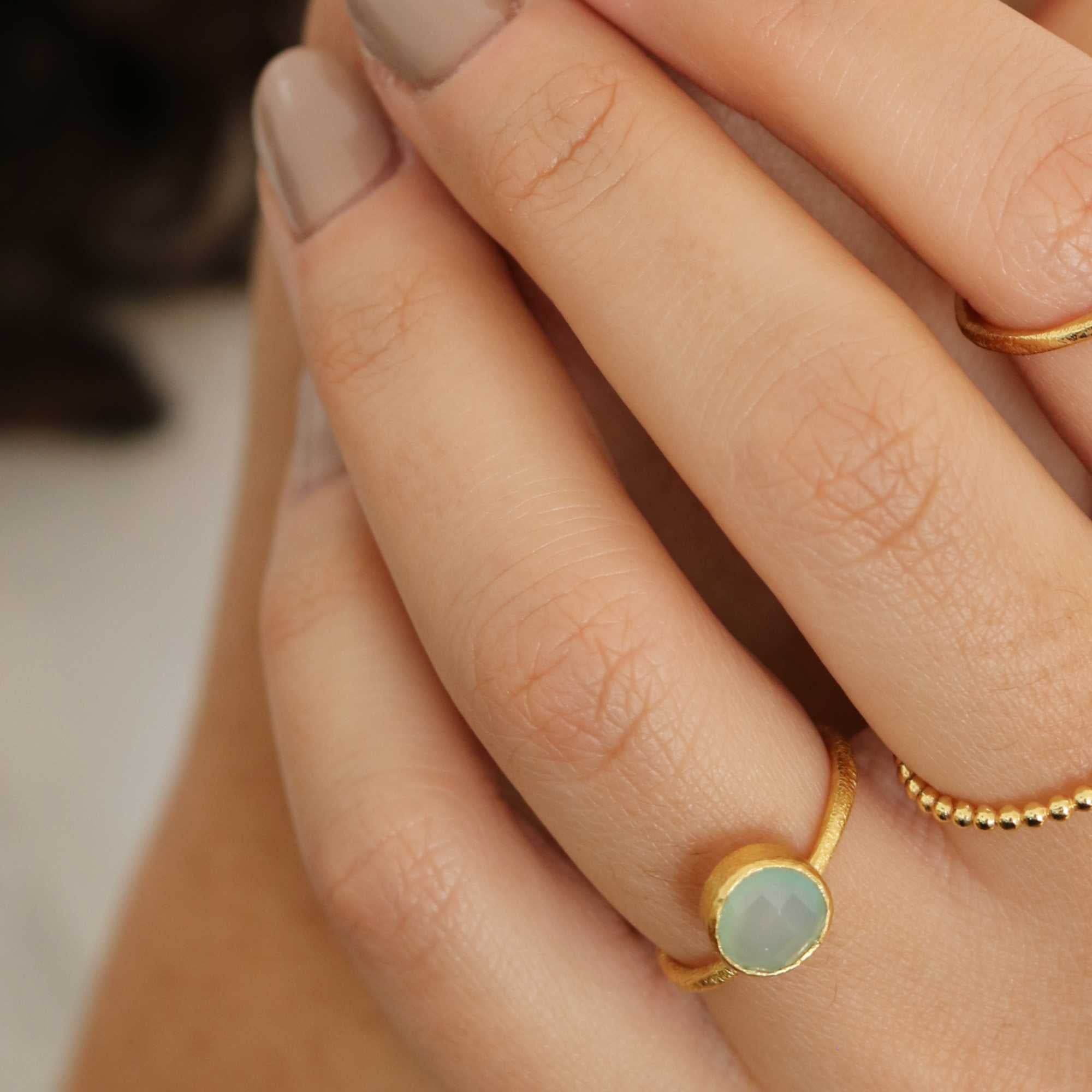 A model showcasing the Dew Drops Oval Aqua Chalcedony Adjustable Ring in gold, featuring a vibrant, bezel-set oval aqua chalcedony gemstone centered on a minimalist textured gold band, styled alongside other delicate stacking rings.