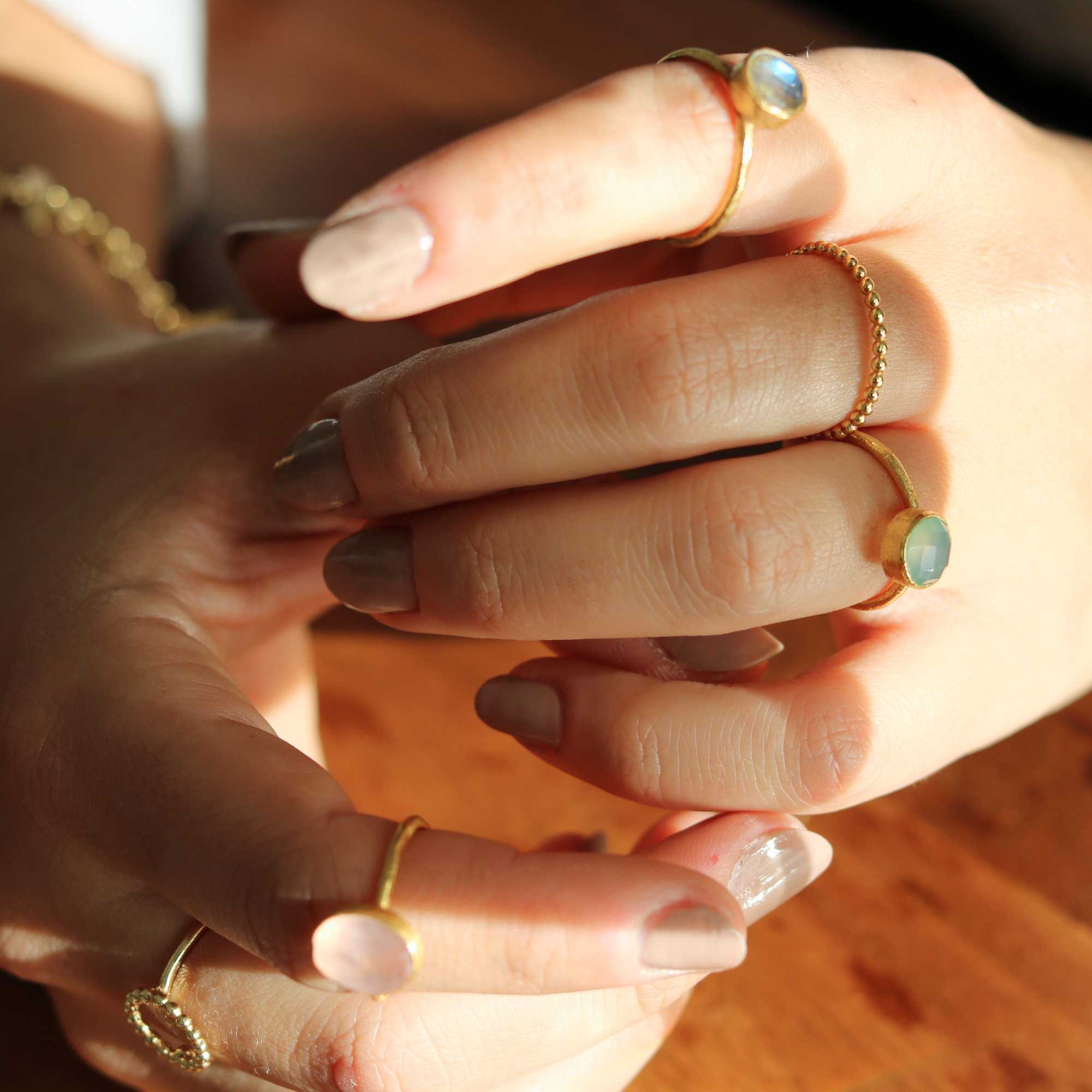 A model showcasing the Dew Drops Oval Aqua Chalcedony Adjustable Ring in gold, featuring a vibrant, bezel-set oval aqua chalcedony gemstone centered on a minimalist textured gold band, styled alongside other delicate stacking rings.