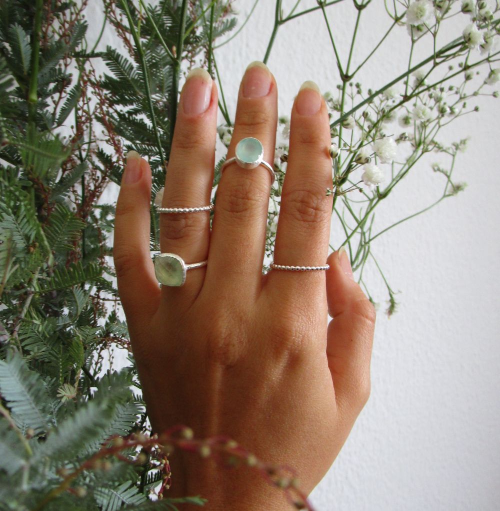 Close-up of a hand wearing a silver oval aqua chalcedony ring with a soft blue-green gemstone and a minimalist polished silver band, photographed against a botanical background.