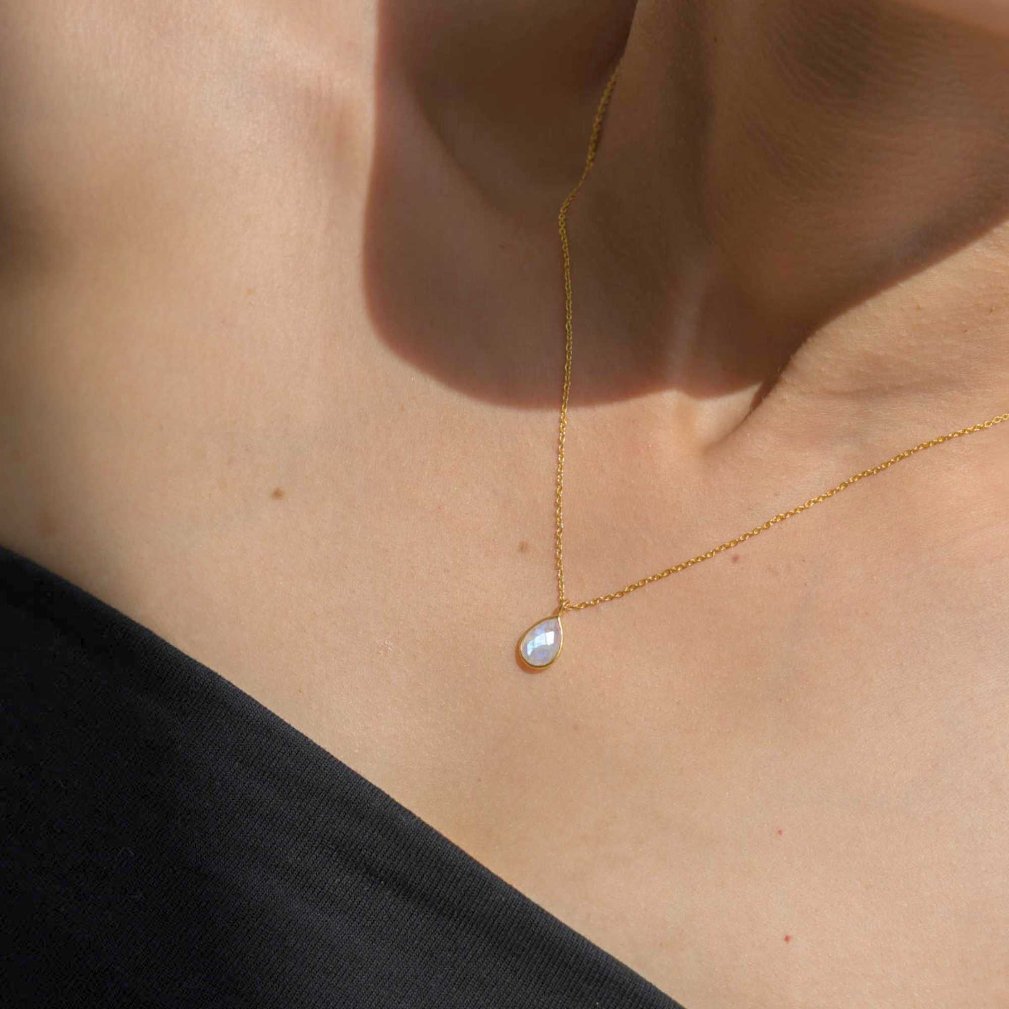 A gold pendant necklace featuring a delicate, bezel-set teardrop rainbow moonstone charm on a fine chain, shown both worn on a model's neckline and flat-layed on a decorative white dish with a mandarin orange and green leaves.