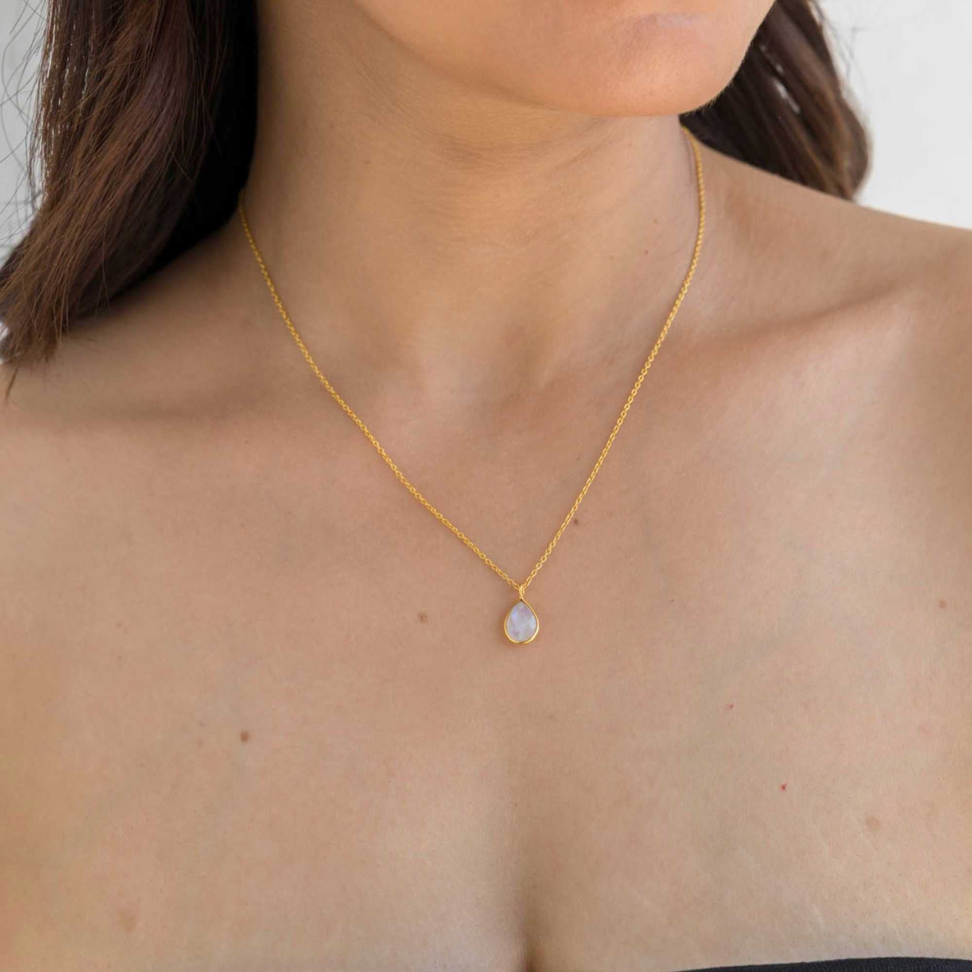 A gold pendant necklace featuring a delicate, bezel-set teardrop rainbow moonstone charm on a fine chain, shown both worn on a model's neckline and flat-layed on a decorative white dish with a mandarin orange and green leaves.