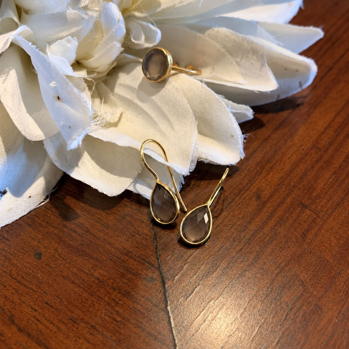 A pair of gold teardrop drop earrings with faceted smoky brown stones, displayed on a white fabric flower prop over a wooden surface.